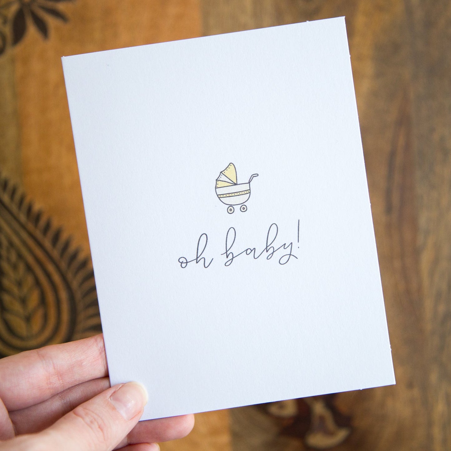 A hand holding a white greeting card with the words "oh baby!" printed below an illustration of a yellow baby stroller