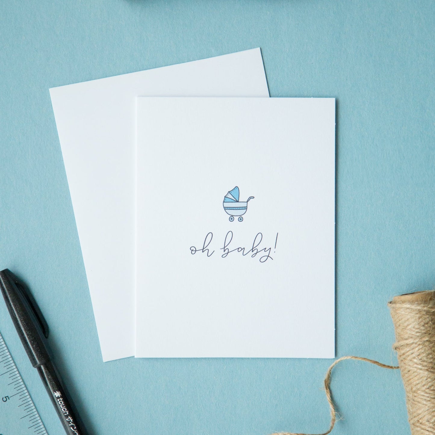 A minimalist baby shower card with a blue baby icon and the text 'oh baby!' on the front, accompanied by a white envelope, lying flat on a blue surface with a ruler, pen, and string arranged beside it.