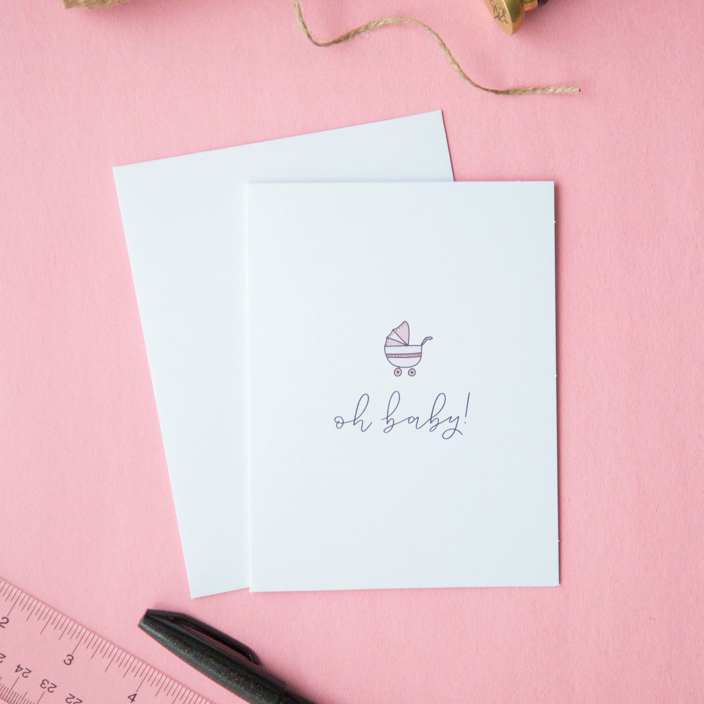 A white A2 sized baby shower card with minimalist design, accompanied by a white envelope, displayed on a pink surface. There is a pen and a ruler alongside, and a small amount of string is visible in the top right corner.