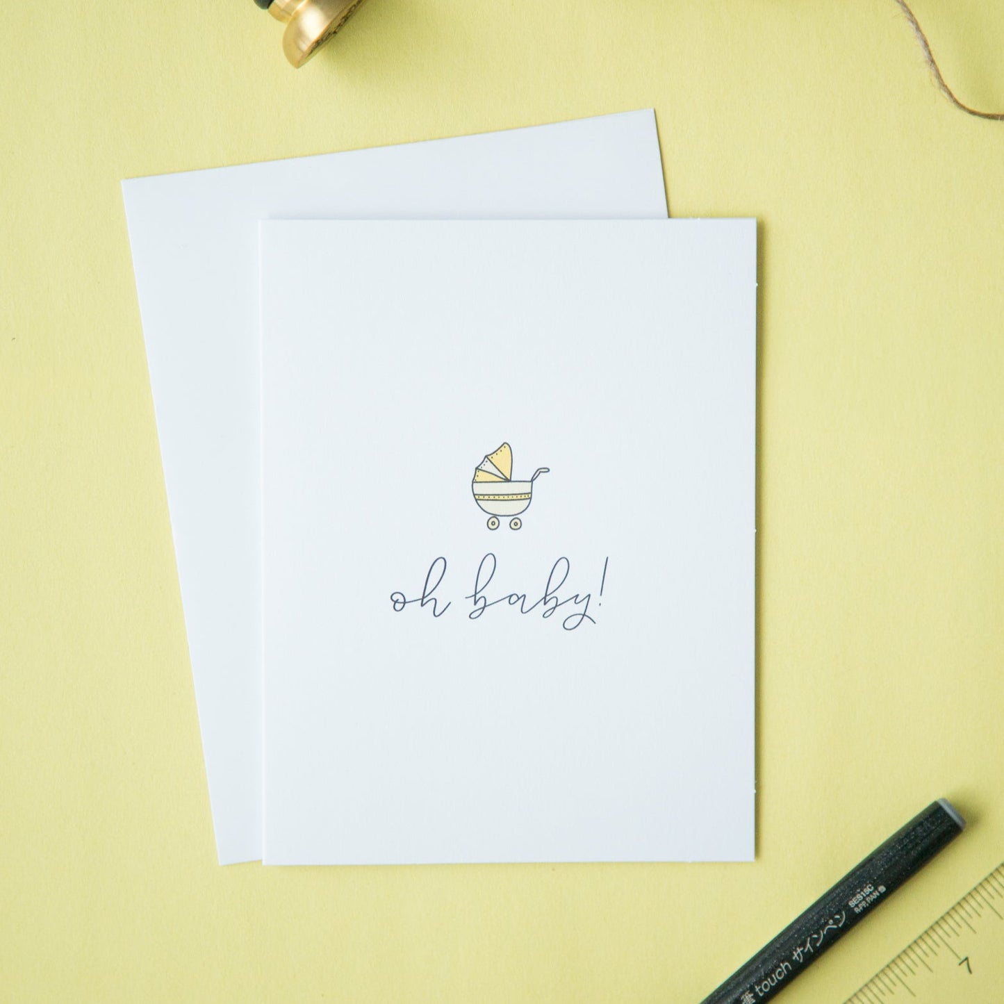 A minimalist style gender neutral baby shower card with the text 'oh baby!' written on it, displayed on a yellow background with an envelope, pen, and ruler.