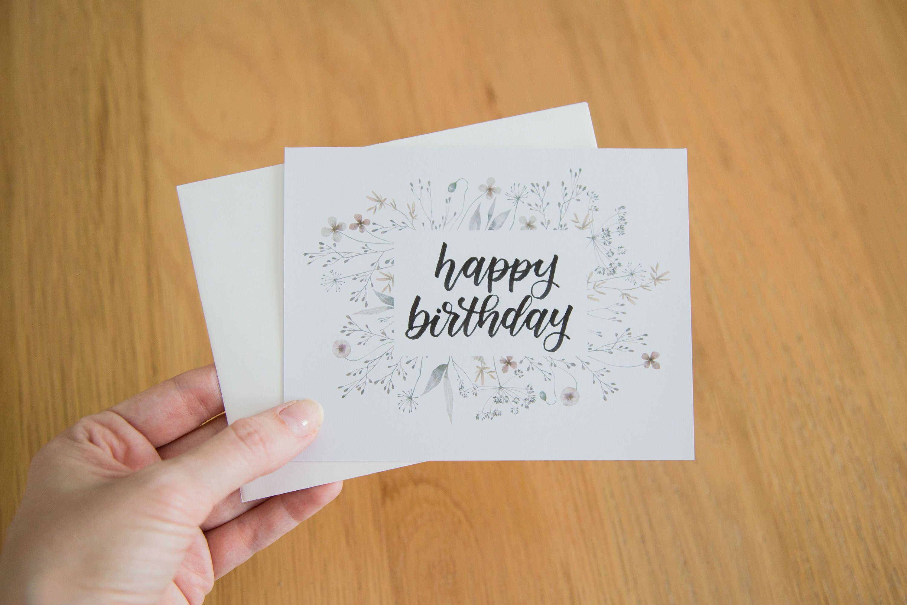 A hand holding a white greeting card with the words "happy birthday" handwritten in black, surrounded by a flower border