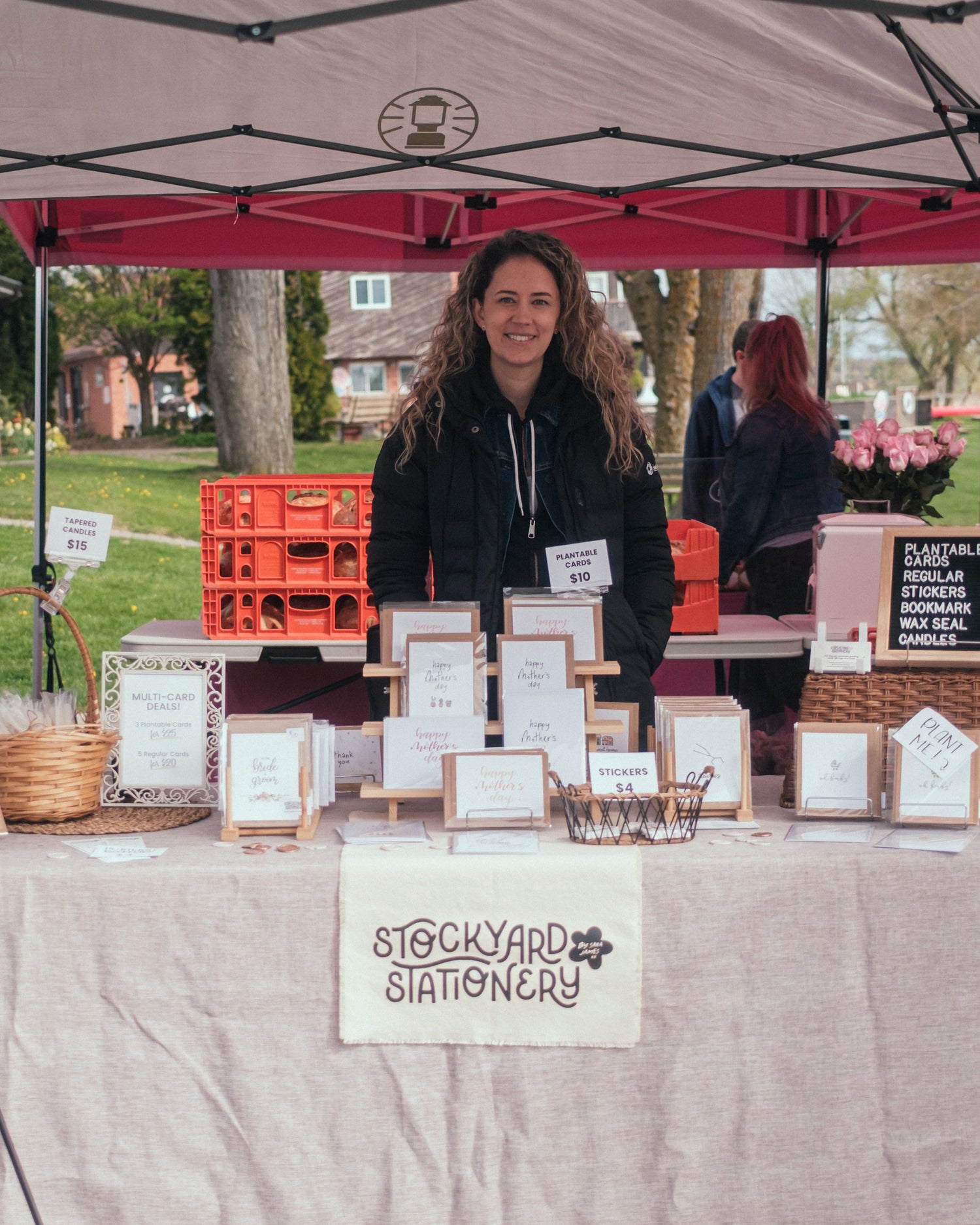 Woman standing behind a table with 'Stockyard Stationery' at an outdoor farmer's market event.