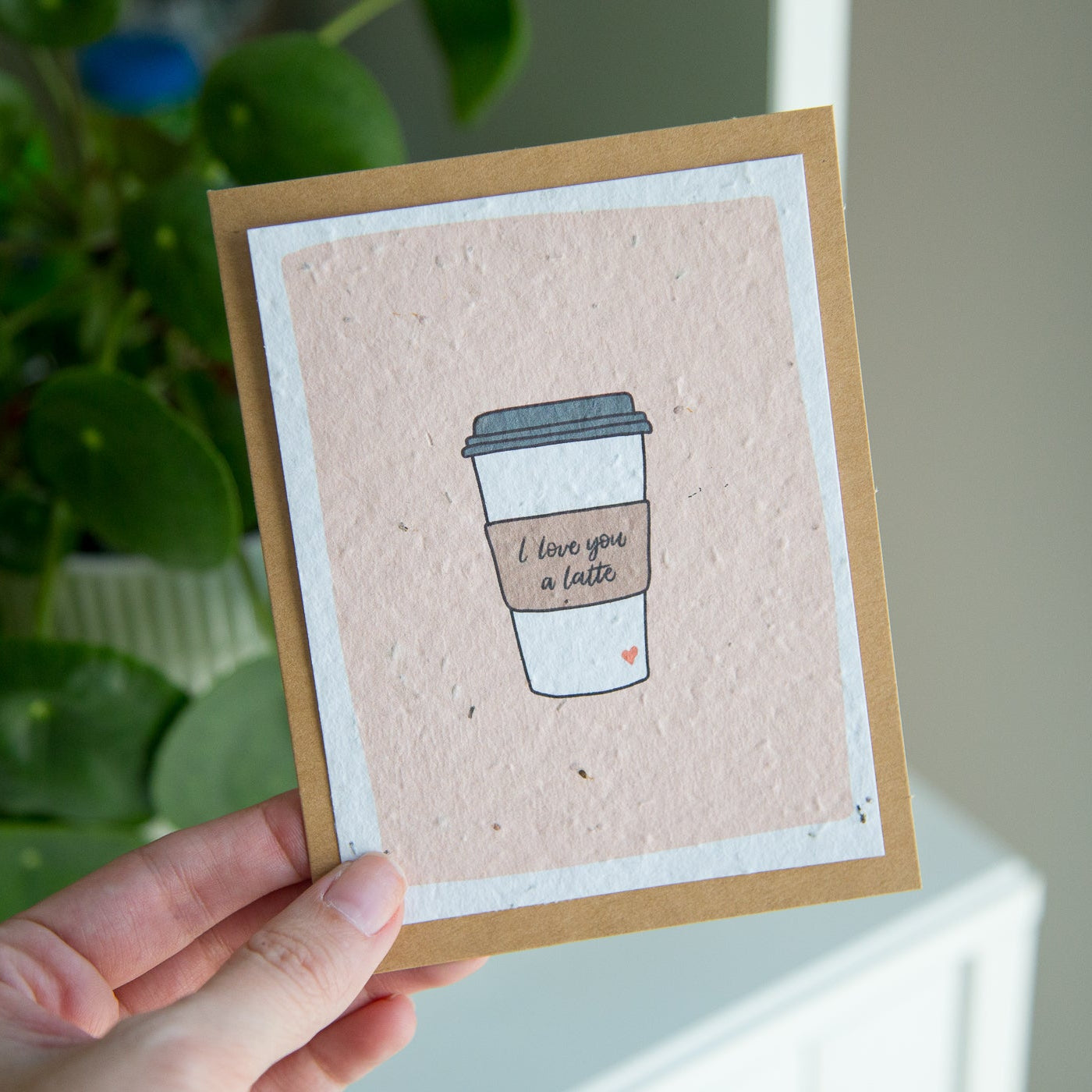 Card with a coffee cup design held by a hand, with a plant in the background