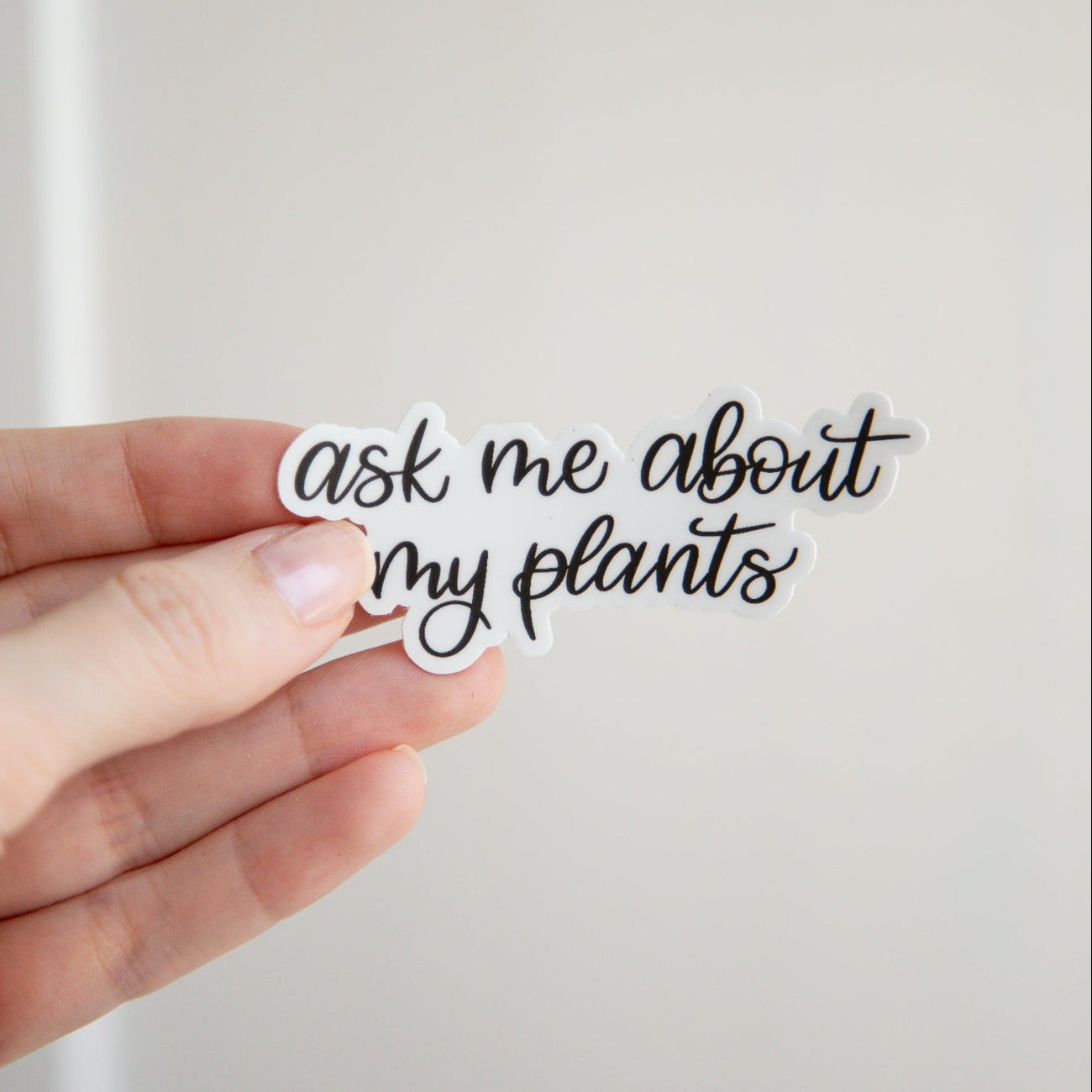 Hand holding a small black and white sticker with 'ask me about my plants' text on a plain background