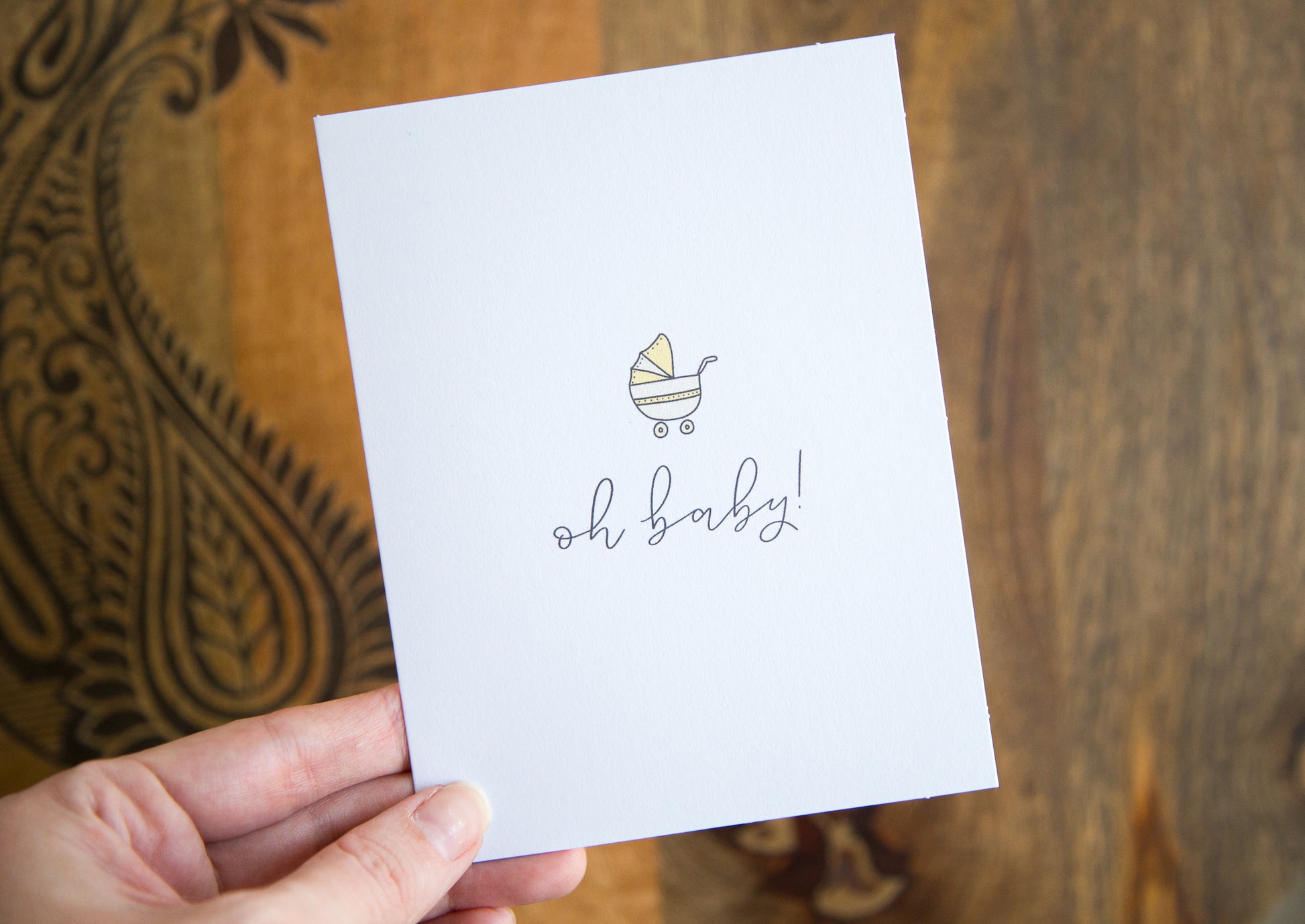 A hand holding a white greeting card with the words "oh baby!" printed below an illustration of a yellow baby stroller