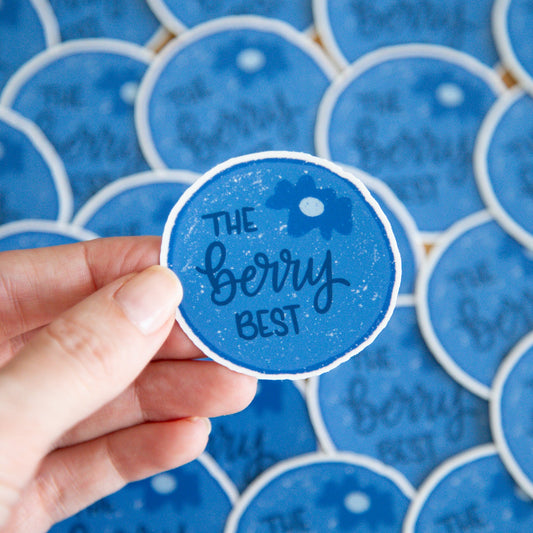 Hand holding a blueberry circular sticker with 'The Berry Best' text against a blue background with similar stickers.