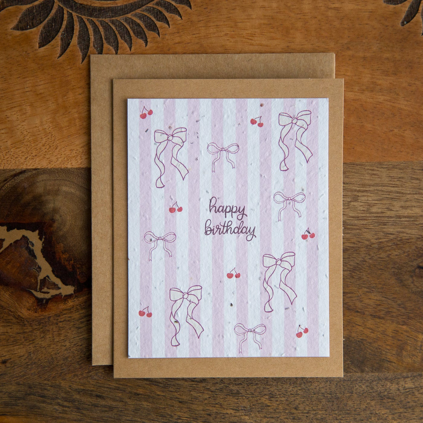 Greeting card with 'Happy Birthday' text, red cherries and pink bow designs on a wooden surface
