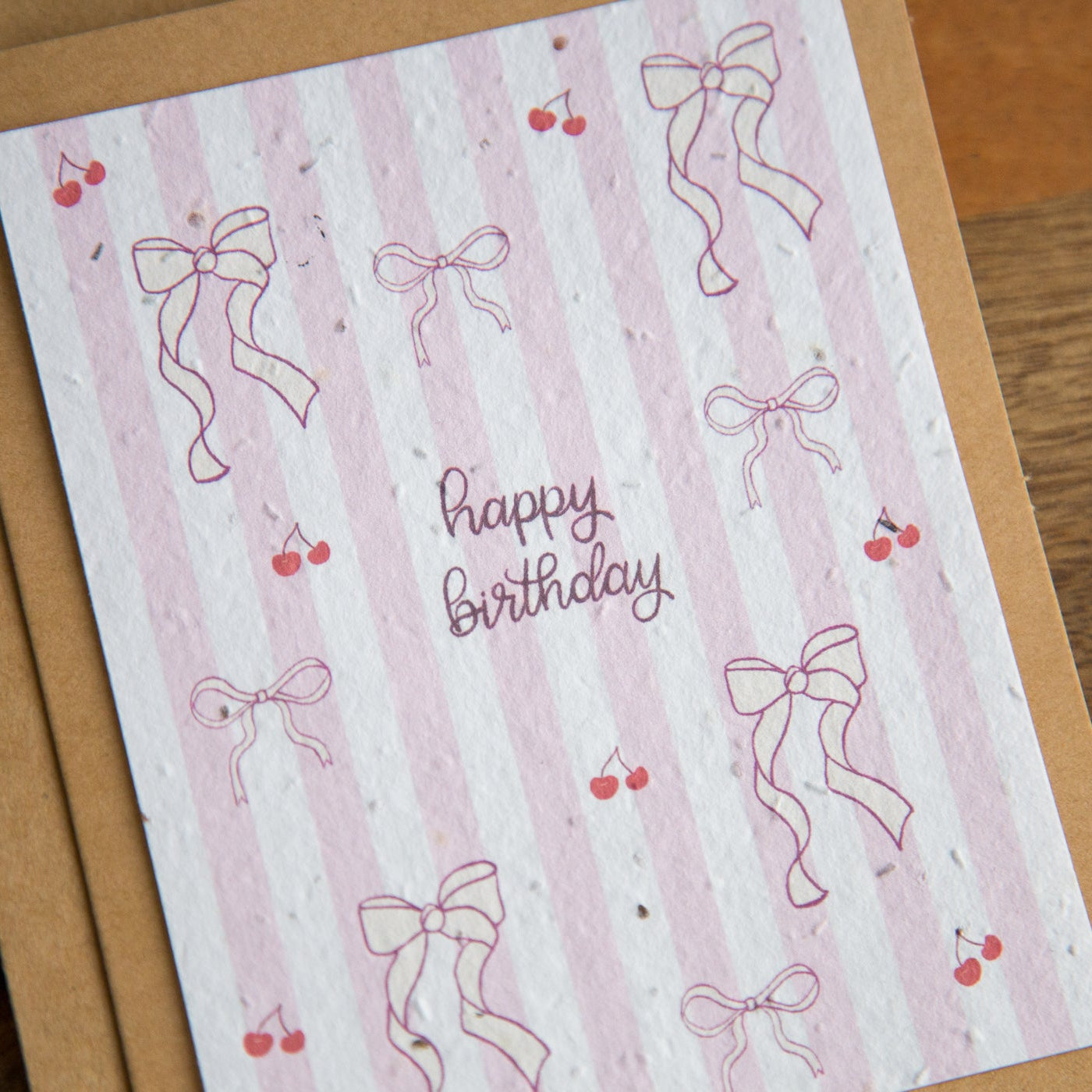 Birthday card with pink bows and cherries on a striped background, placed on a wooden surface.
