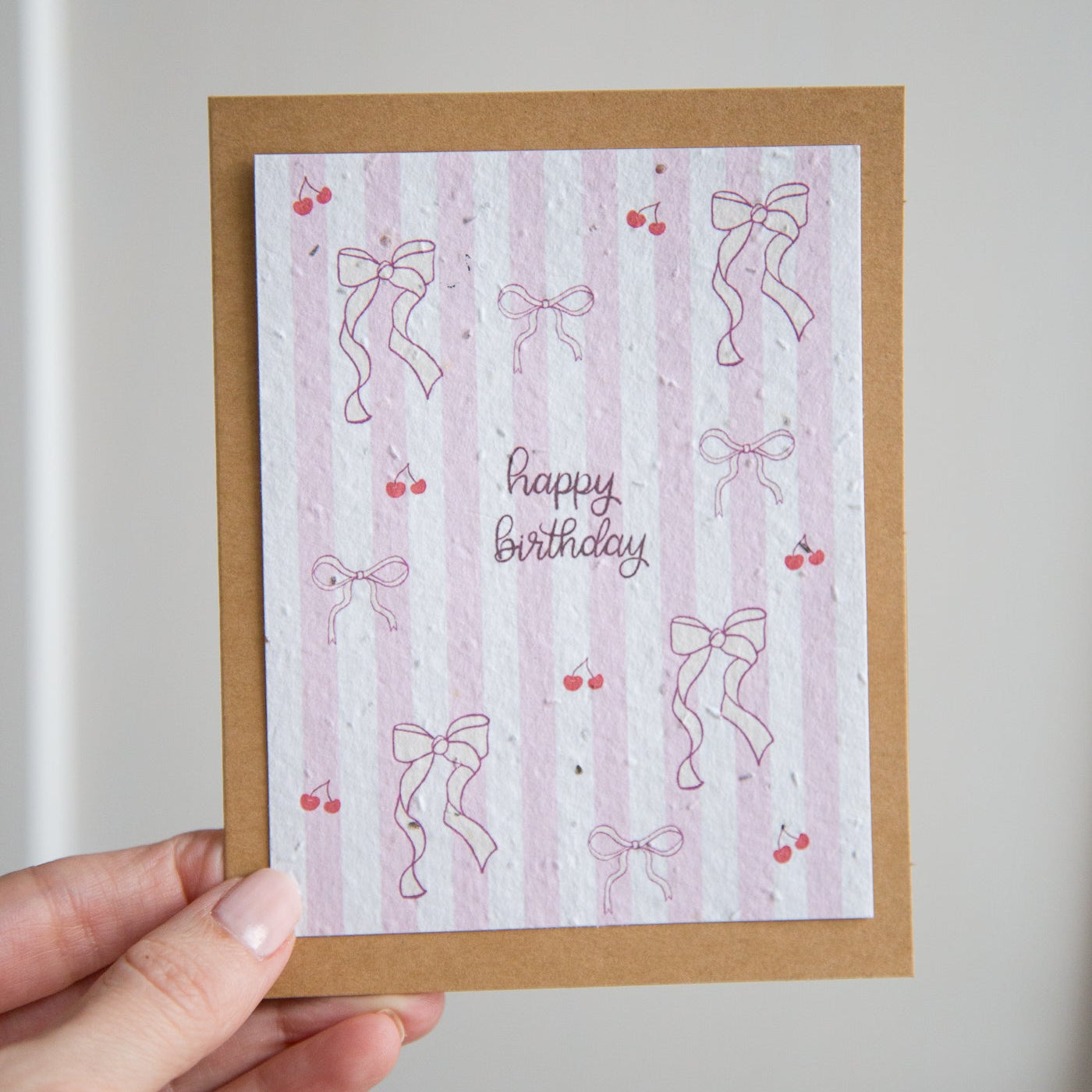 Hand holding a birthday card with pink stripes and bows on a plain background