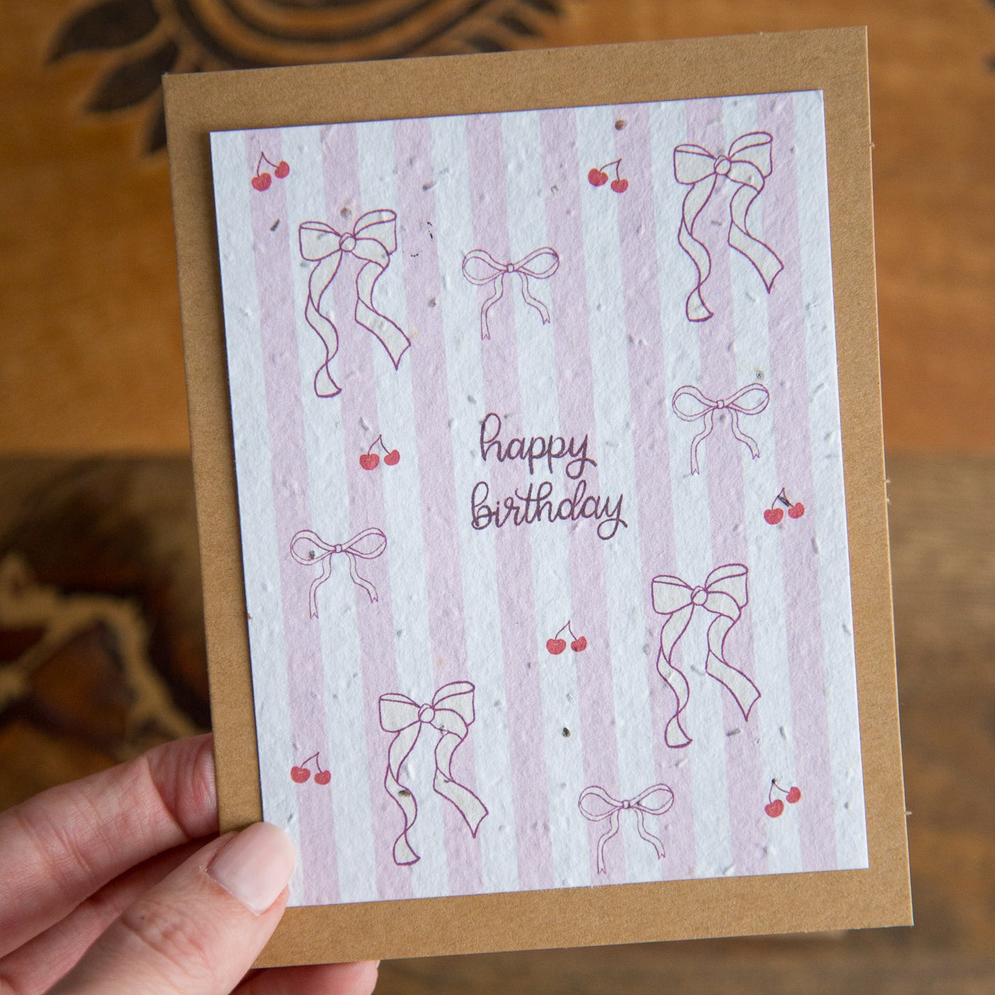 Hand holding a birthday card with pink bows and cherries on a wooden surface