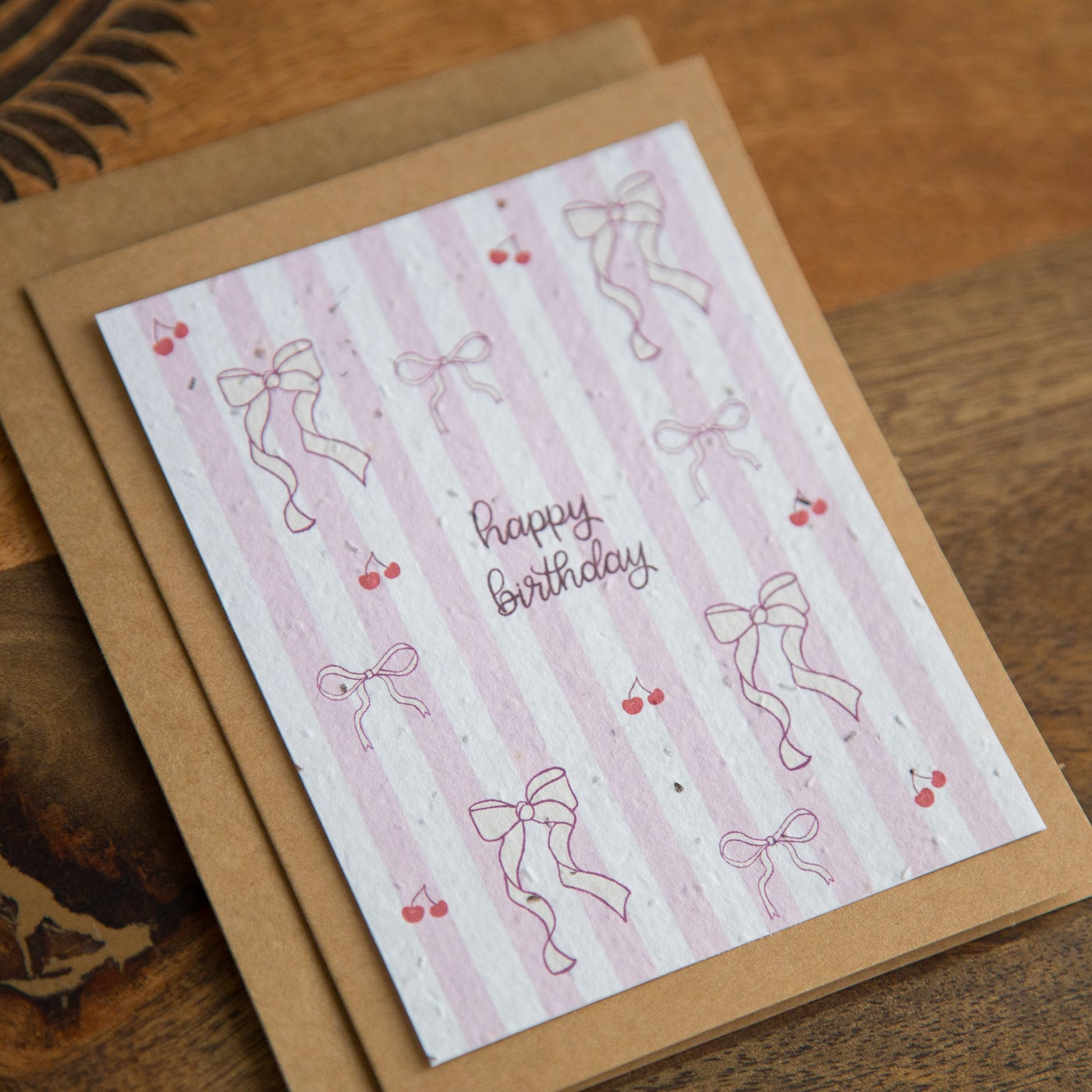 Greeting card with 'Happy Birthday' text and bow designs on a striped background, placed on a wooden surface.