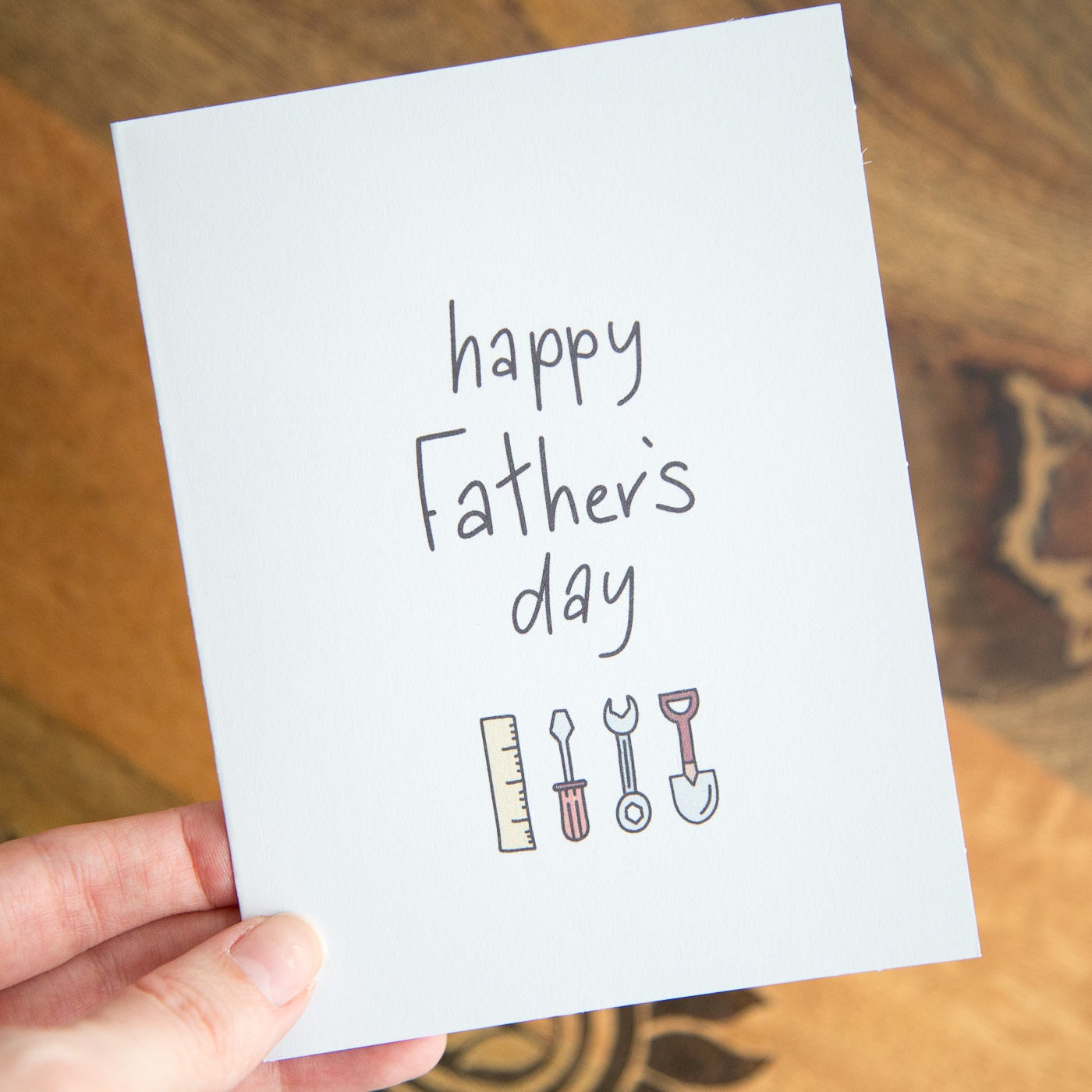 Hand holding a Father's Day card with tools illustration on a wooden surface