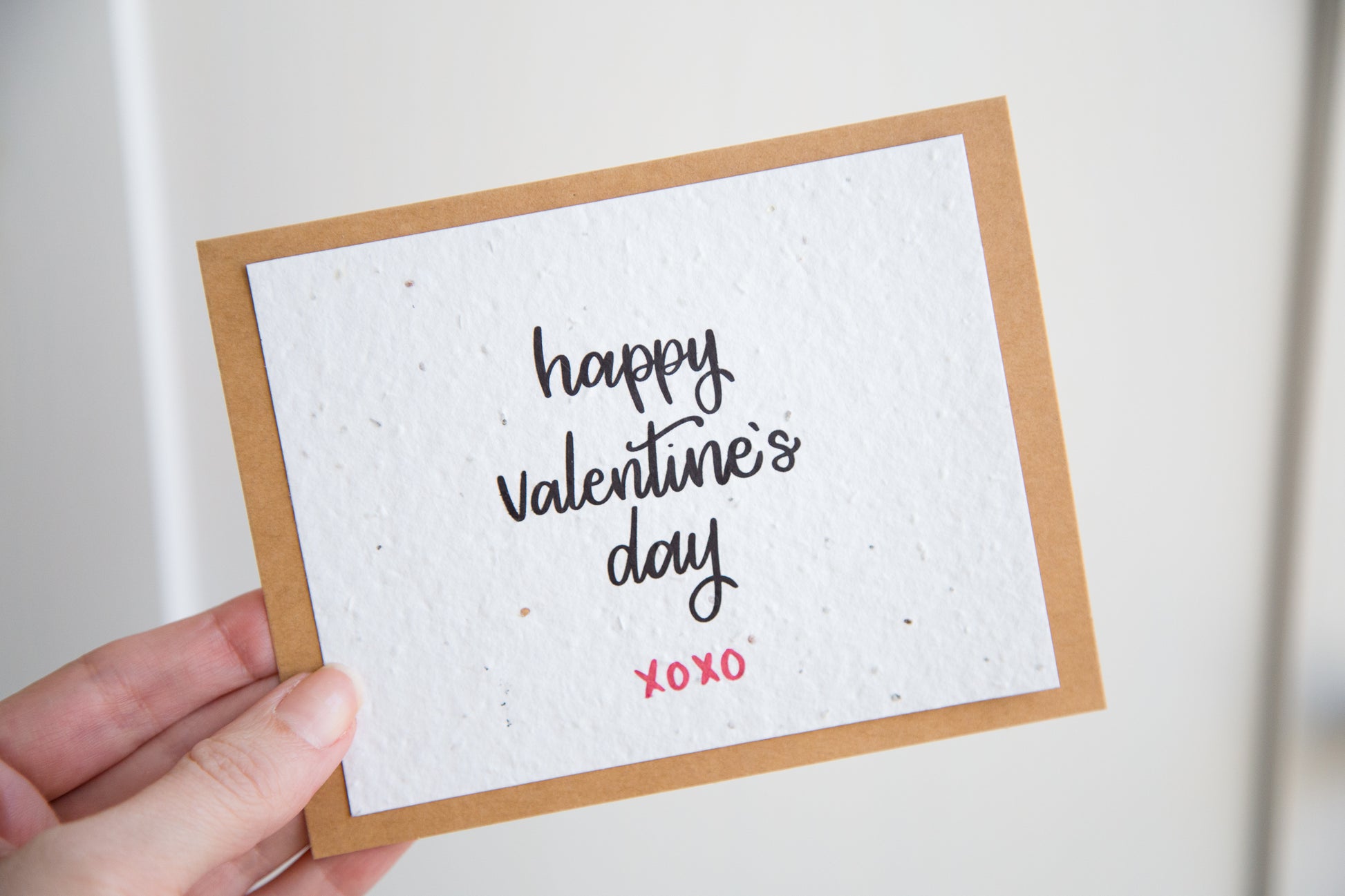 A hand holding a plantable greeting card with the words "happy Valentine's Day xoxo" printed in calligraphy in black on white paper