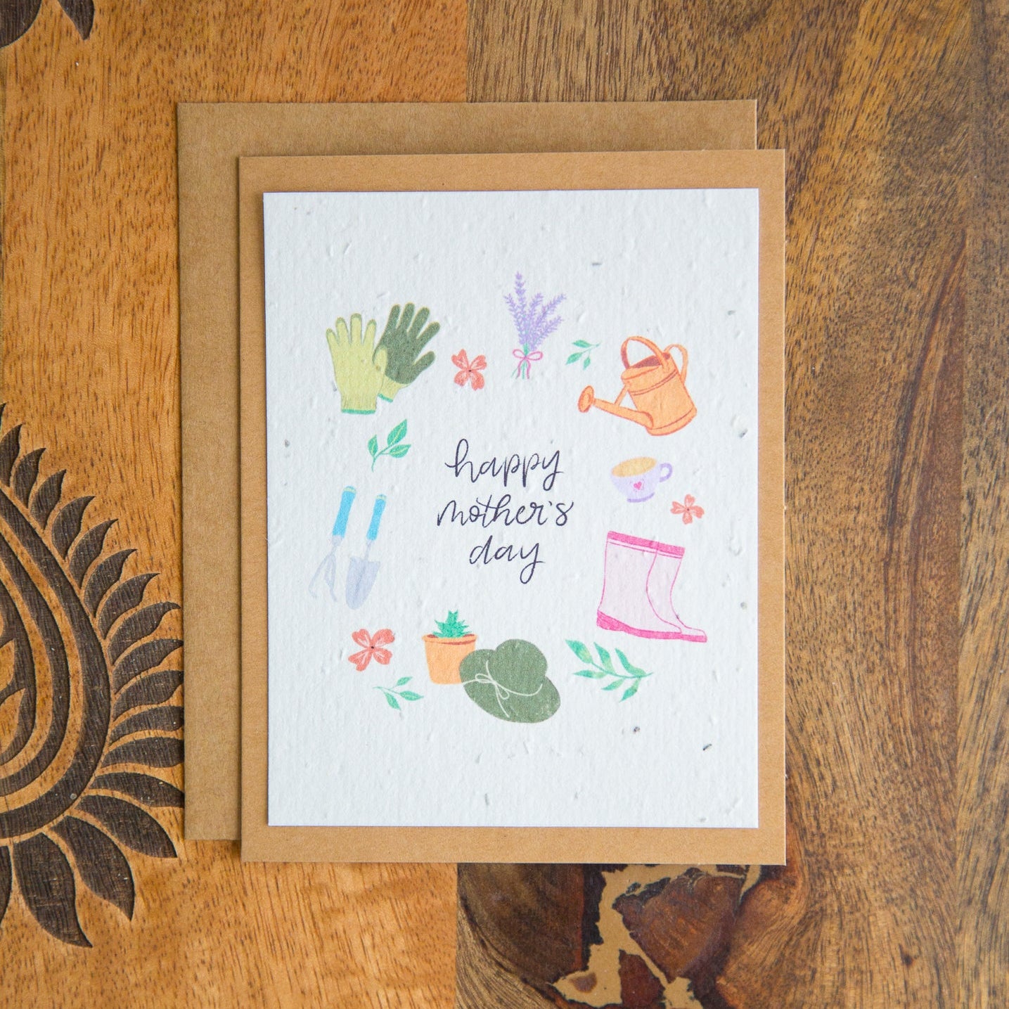 Greeting card with 'Happy Mother's Day' text and gardening-themed illustrations on a wooden surface.