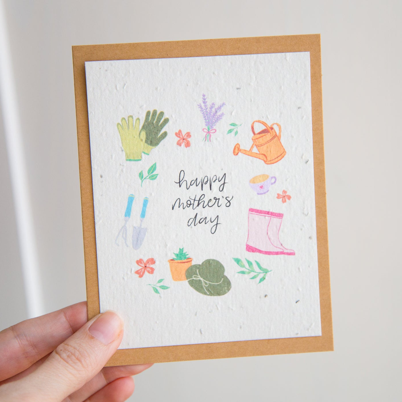 Hand holding a Mother's Day card with gardening-themed illustrations on a plain background