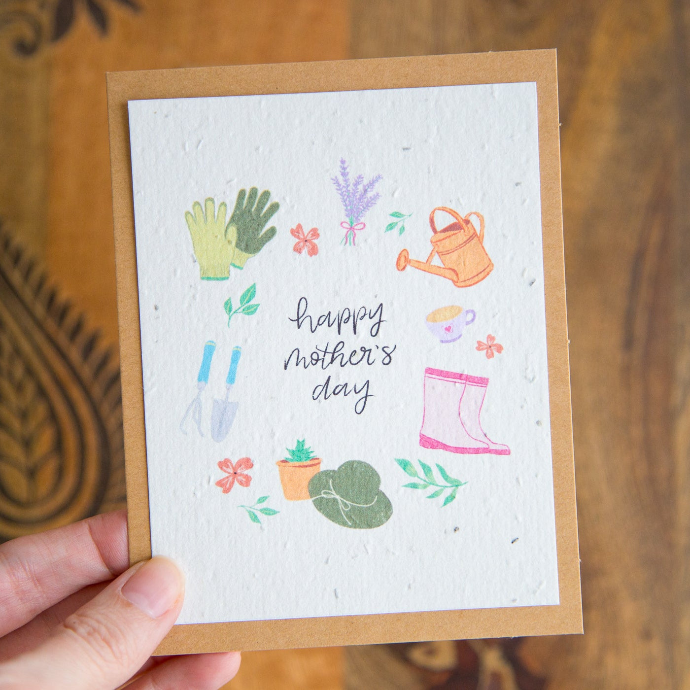 Hand holding a Mother's Day card with gardening-themed illustrations on a wooden surface.