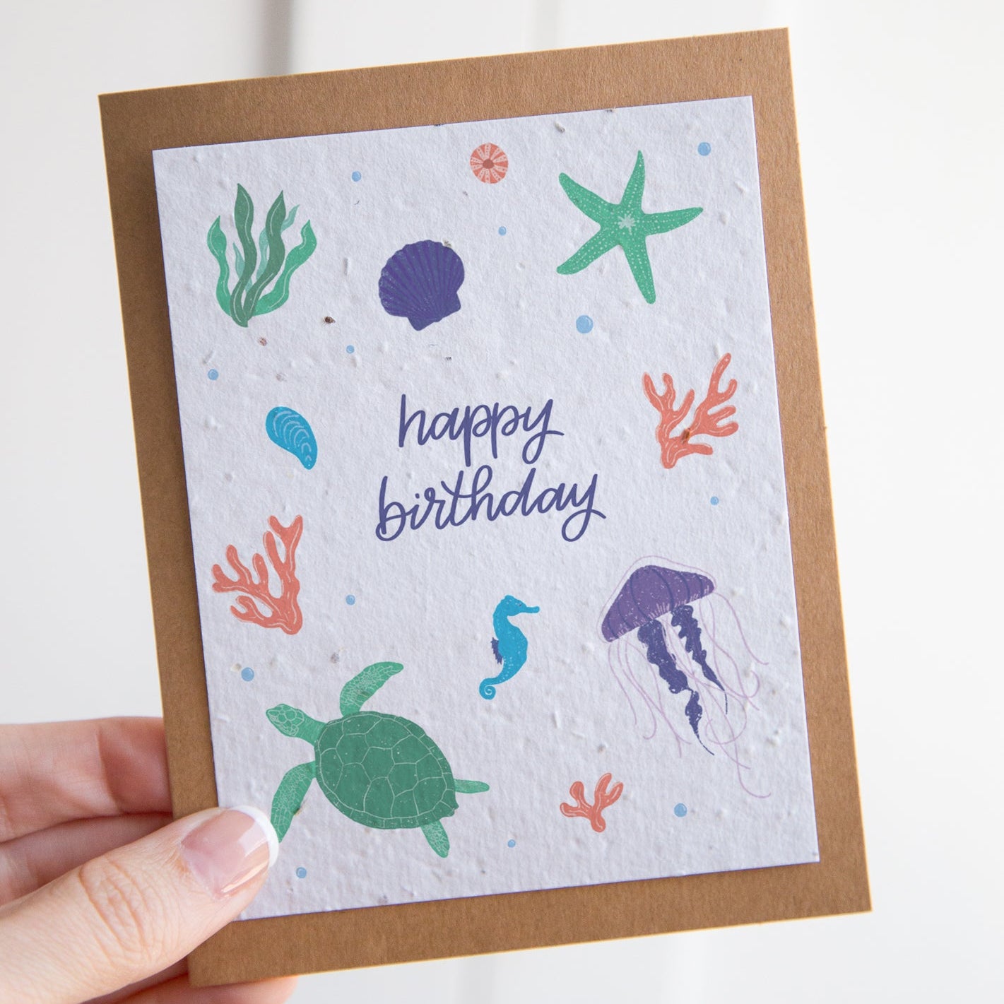 A handmade square birthday card with an ocean theme, held by a hand against a white background. The card has a brown kraft paper border and a white textured center featuring colorful sea life illustrations including a green turtle, starfish, jellyfish, and coral, with 'happy birthday' written in blue script.