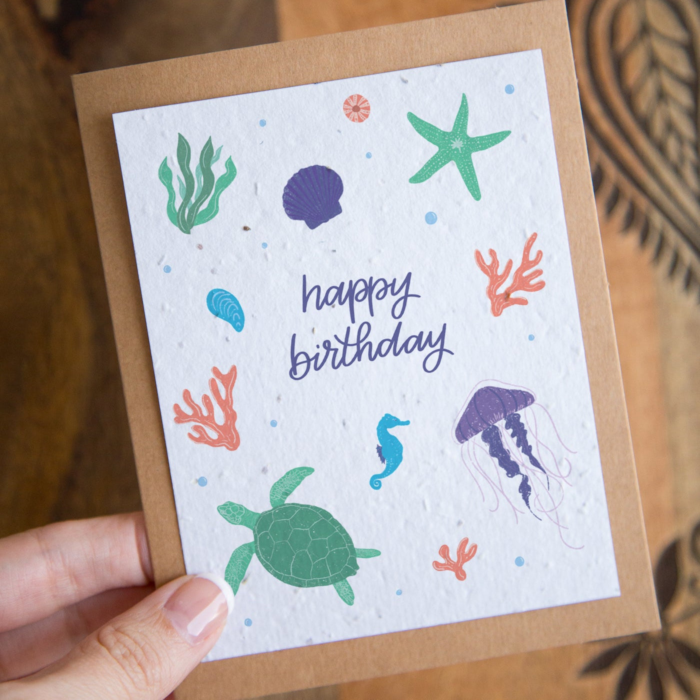 A handmade paper card with an ocean-themed design, held by a hand against a wooden background. The card features a white textured center with illustrations of a green sea turtle, purple jellyfish, green starfish, and coral, alongside the text 'happy birthday' in purple script.