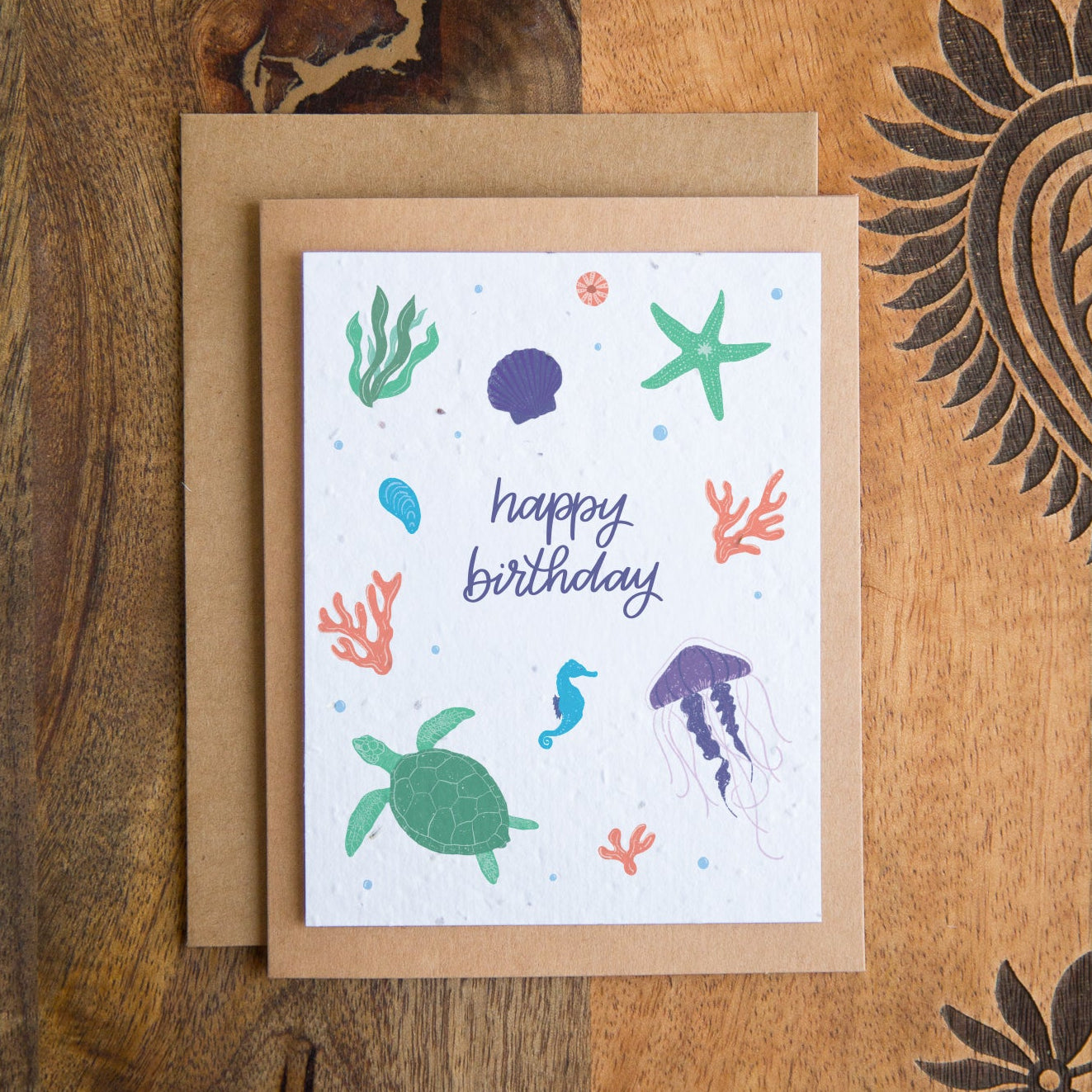 A square birthday card with an underwater theme, featuring illustrations of a green sea turtle, blue jellyfish, green starfish, and coral on a white background with the words 'happy birthday' in blue script. The card is shown with a brown kraft envelope on a rustic wooden background.