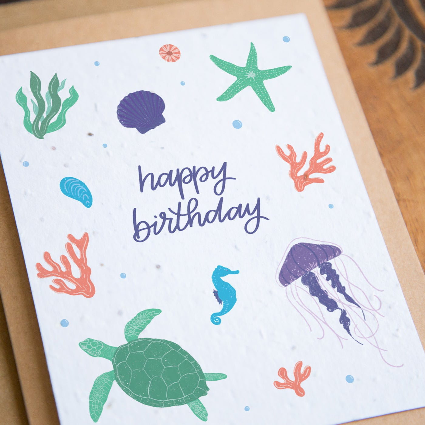 A square white birthday card with colorful ocean-themed illustrations including a green sea turtle, purple jellyfish, blue seahorse, green starfish, coral, and seaweed, with the words 'happy birthday' in purple script. The card is placed on a brown kraft envelope over a wooden surface.