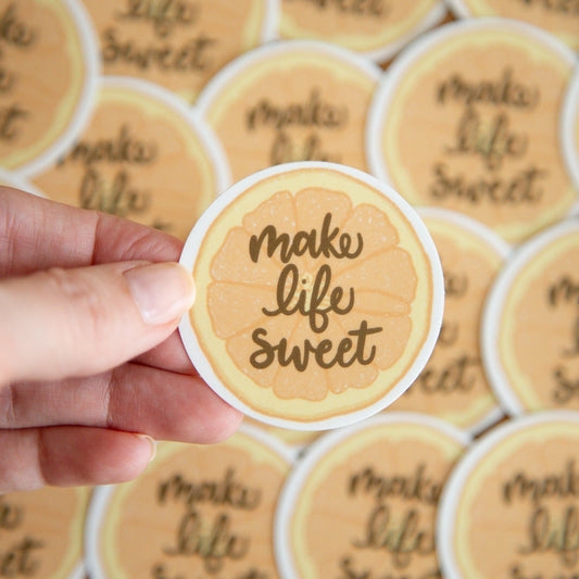 Hand holding a round sticker of an orange slice with 'make life sweet' text against a background of similar stickers.