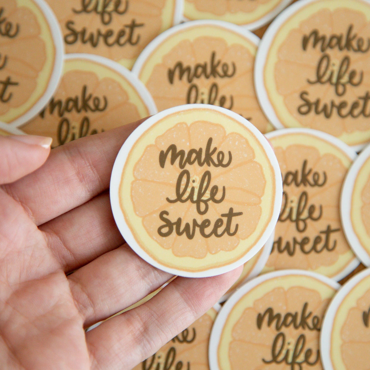 Hand holding a round sticker of an orange slice with 'make life sweet' text against a background of similar stickers
