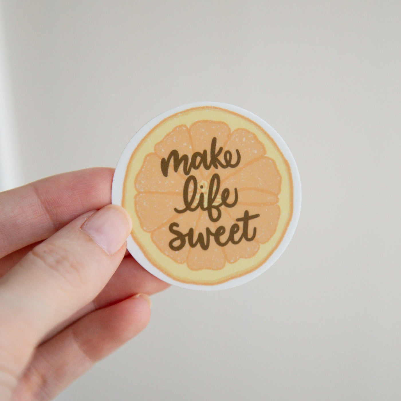 Round orange slice sticker with 'make life sweet' text held by a hand against a light background