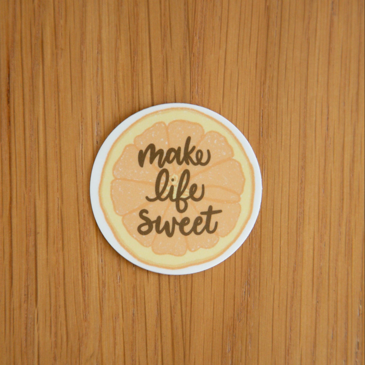 Round sticker of an orange slice with 'make life sweet' text against a wood background.