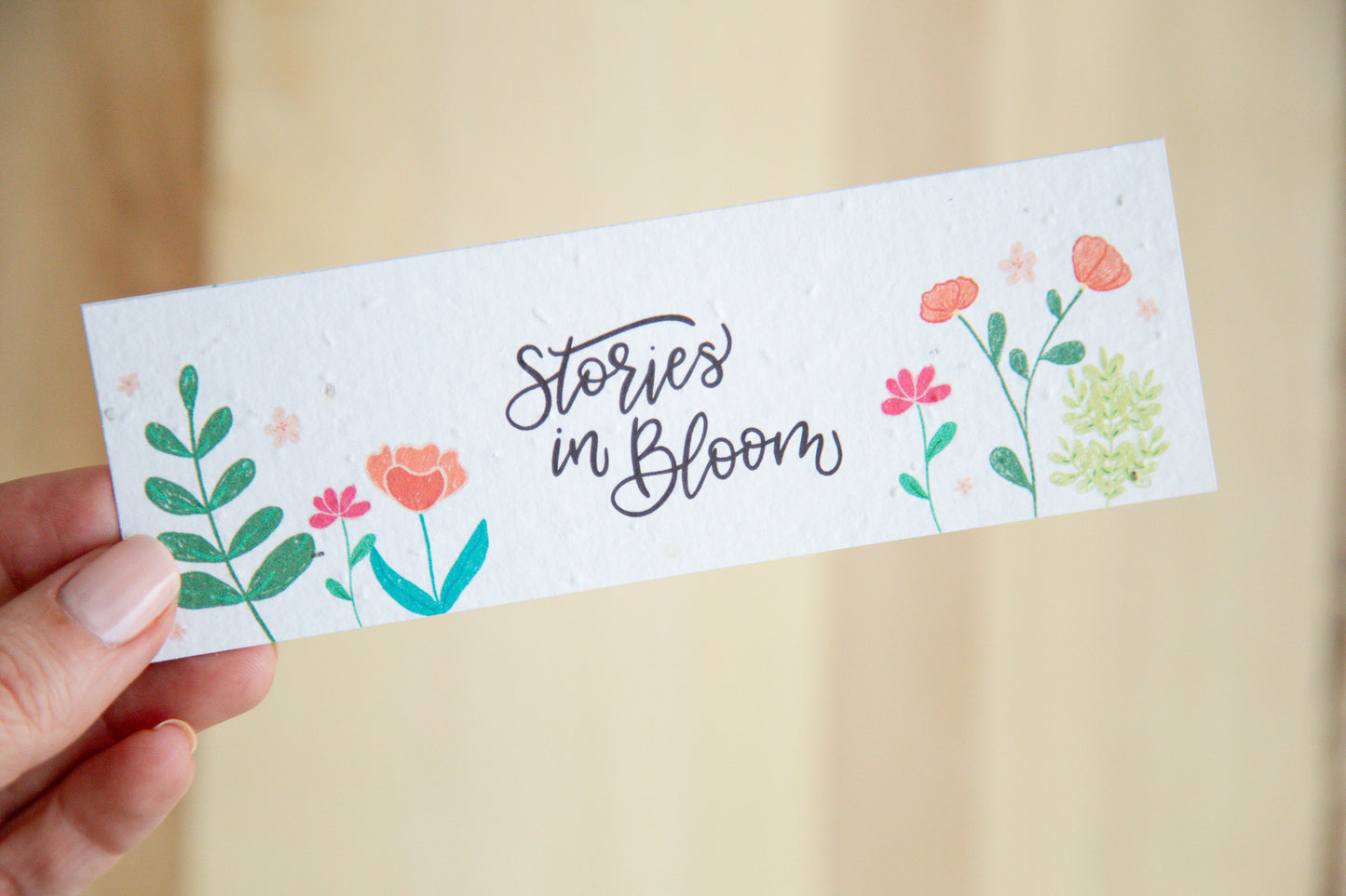 Bookmark with 'Stories in Bloom' text and floral designs held by a hand on a wooden background