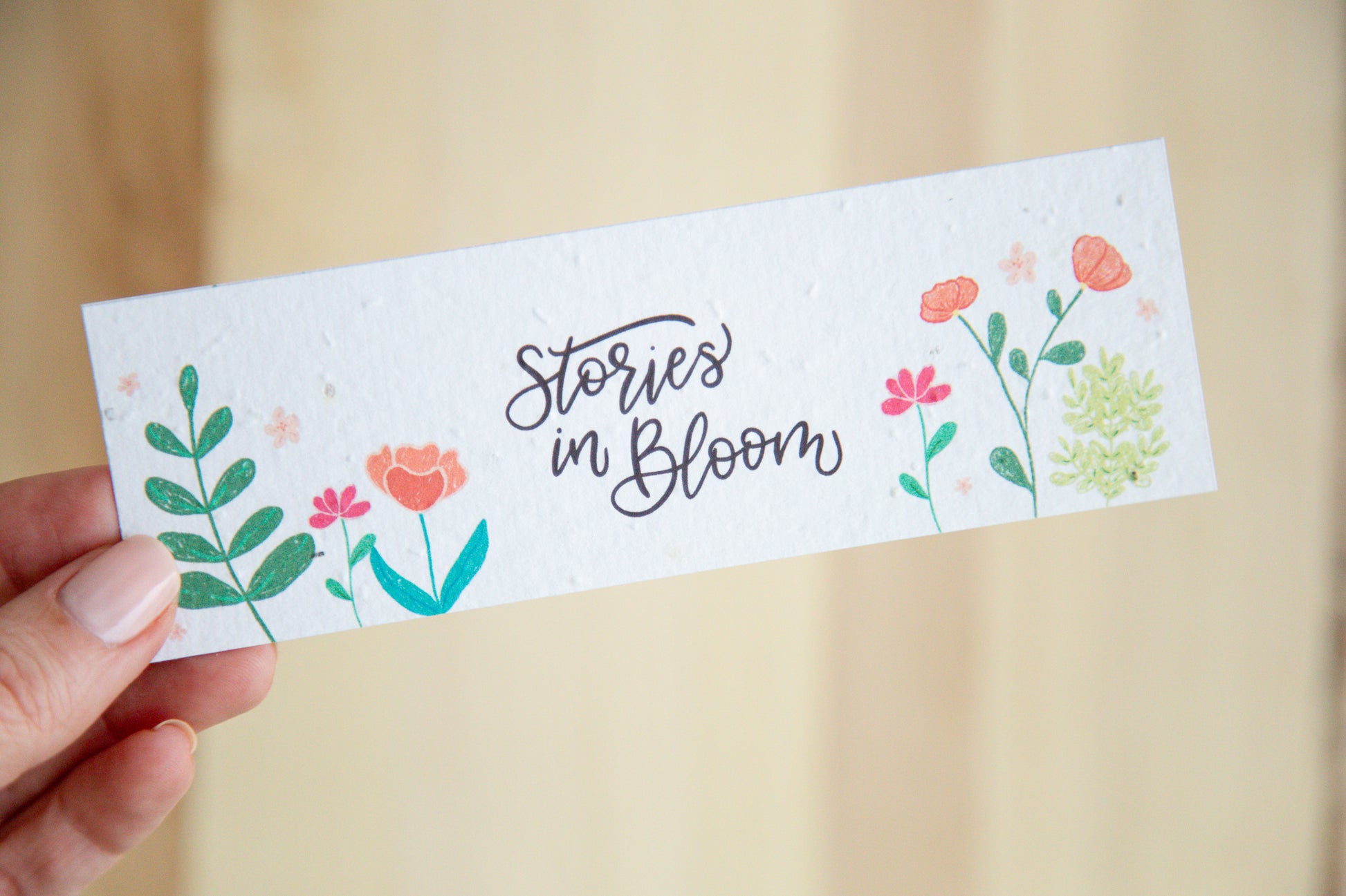 Bookmark with 'Stories in Bloom' text and floral designs held by a hand on a wooden background