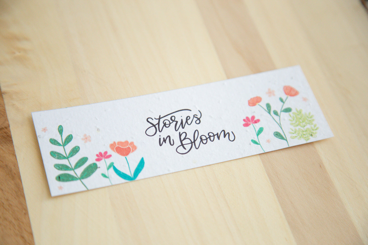 Bookmark with floral design and 'Stories in Bloom' text on a wooden surface