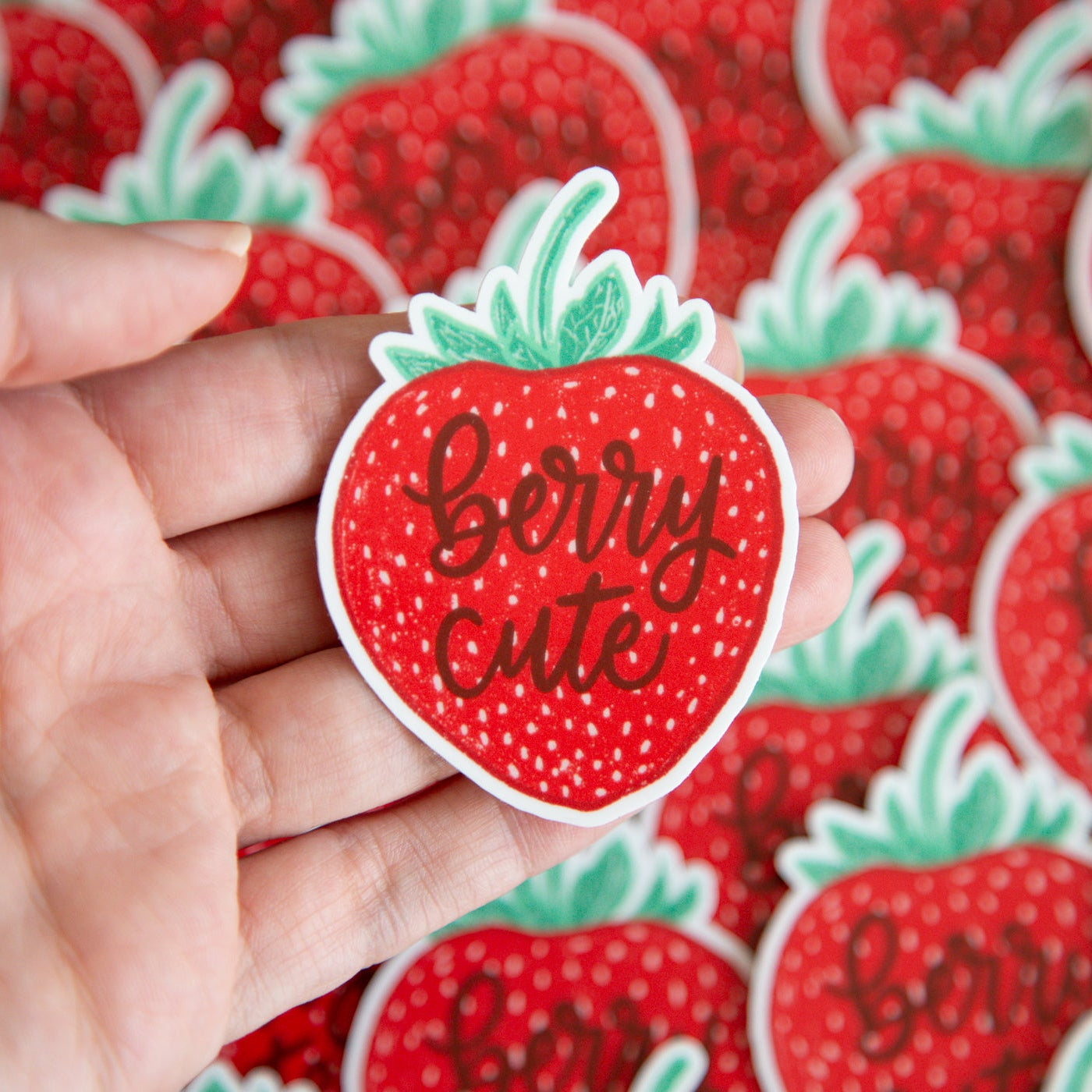 Hand holding a strawberry-shaped sticker with 'berry cute' text against a background of similar stickers.