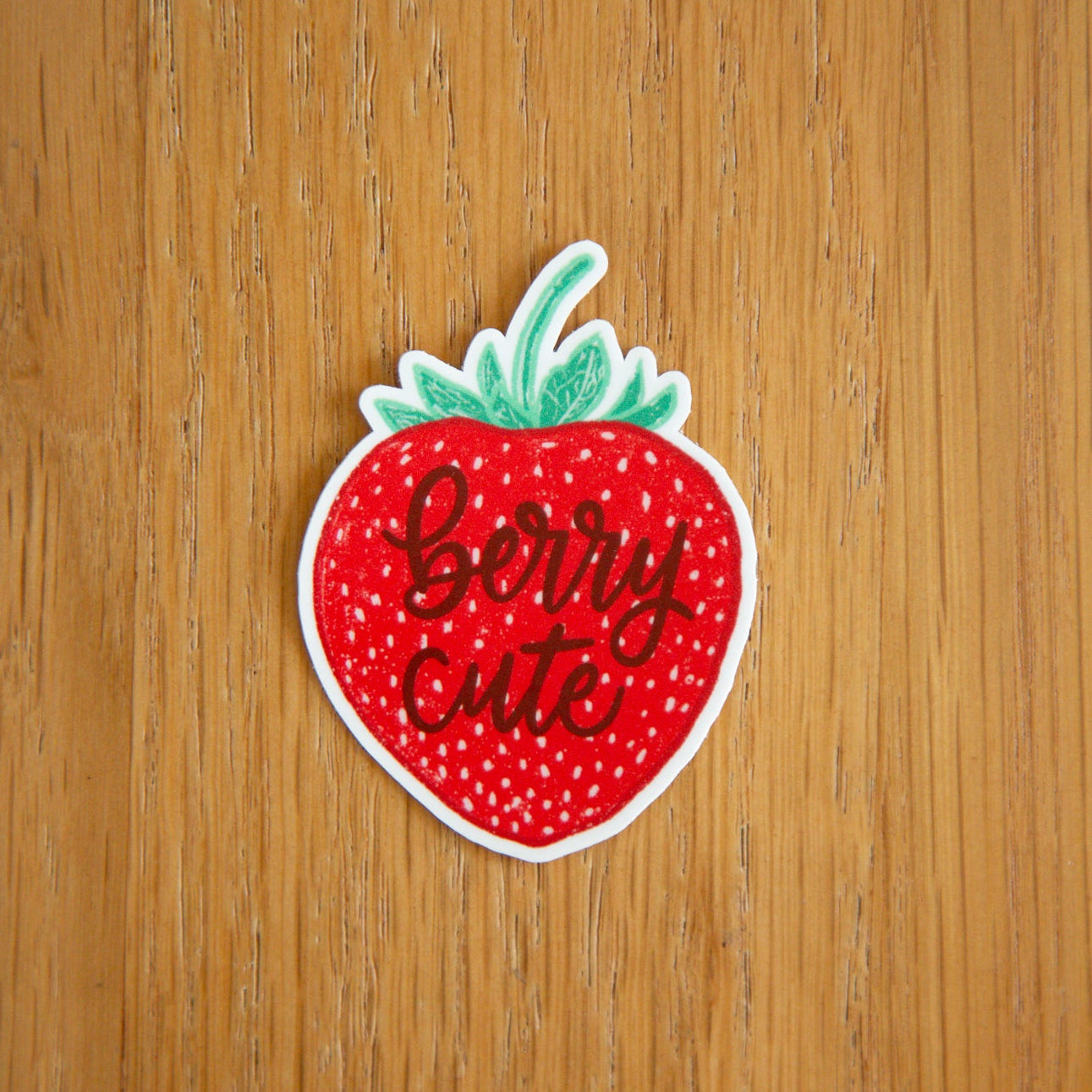 Strawberry-shaped sticker with 'Berry cute' text on a wooden background