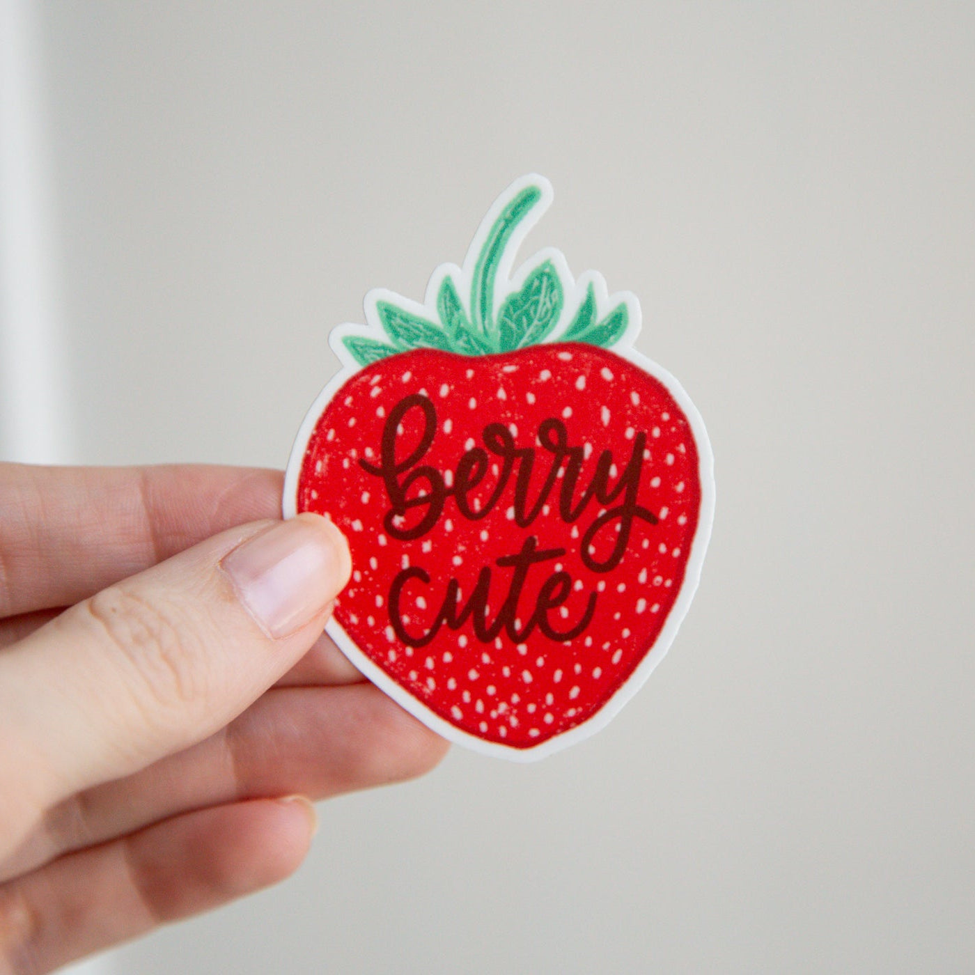 Strawberry-shaped sticker with 'Berry Cute' text held by a hand against a plain background