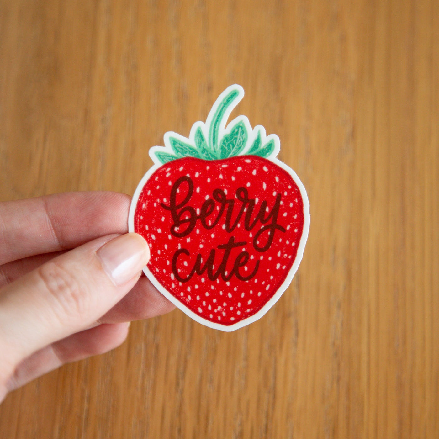 Strawberry-shaped sticker with 'Berry cute' text held by a hand on a wooden surface