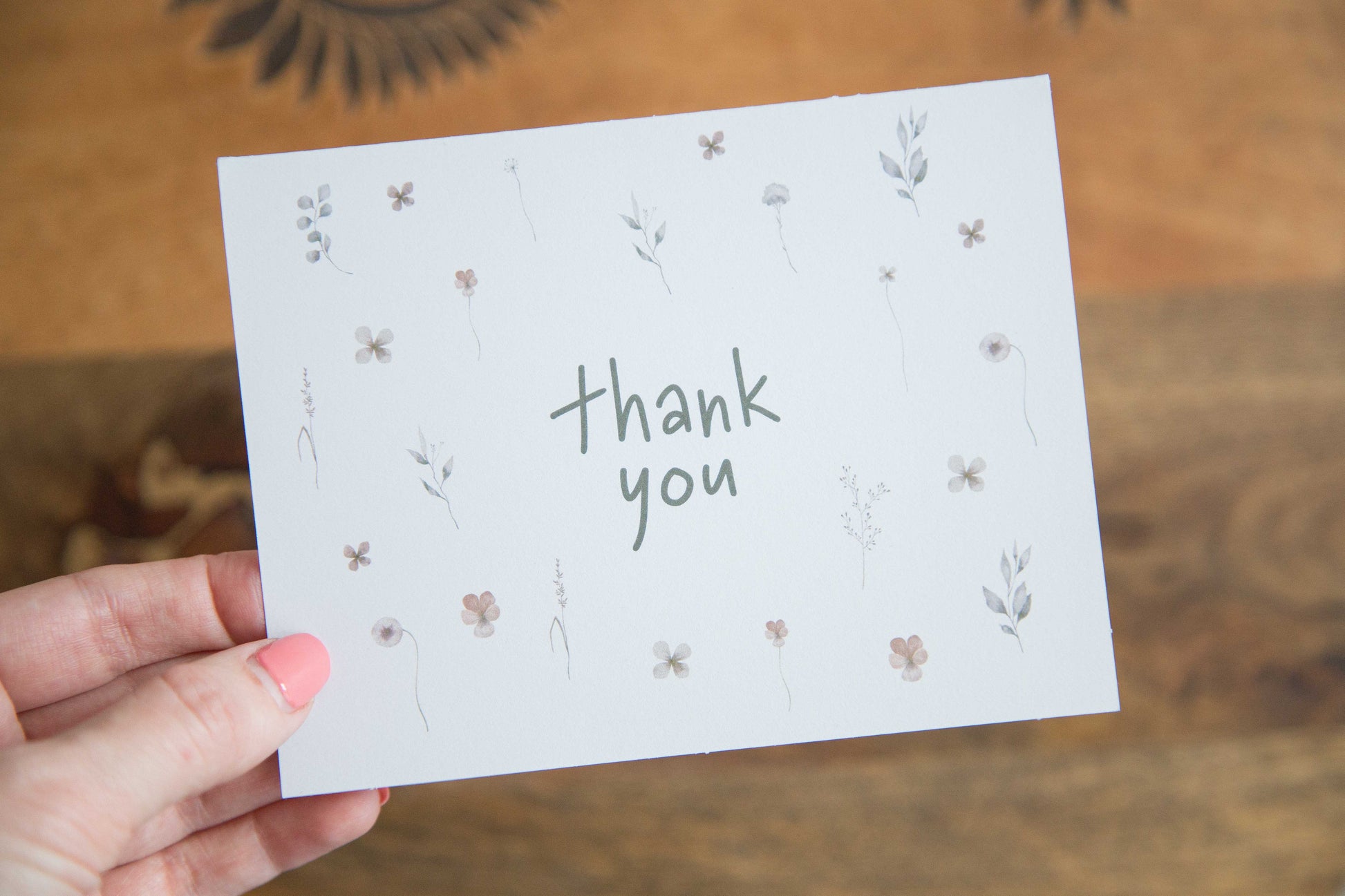 a hand holding a white greeting card with the works "thank you" printed surrounded by watercolour images of flowers