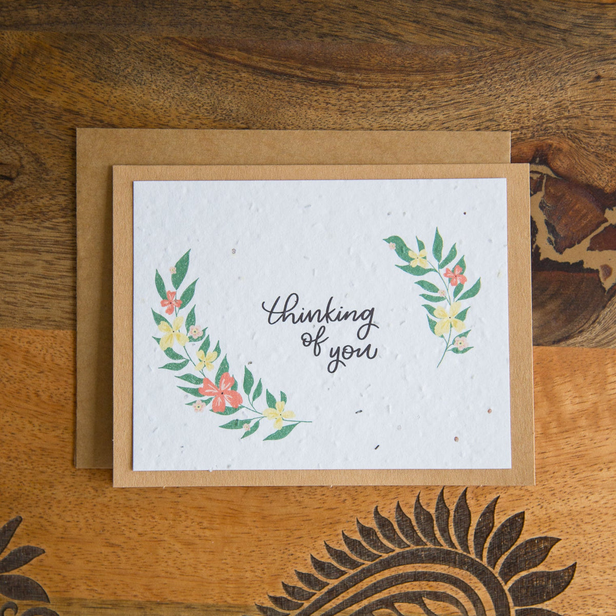 A square white greeting card with a kraft envelope, featuring the words 'thinking of you' in black script and watercolor floral wreaths in green, pink, and yellow, displayed on a wooden surface with a carved paisley design.