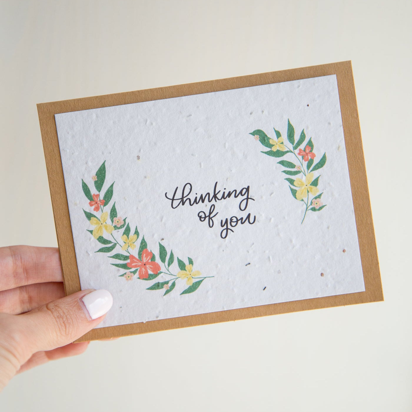 A handmade greeting card with a kraft paper border and a textured white center featuring the phrase 'thinking of you' in black script, framed by green leaves and small pink flowers. A hand with white nail polish holds the card against a plain off-white background.