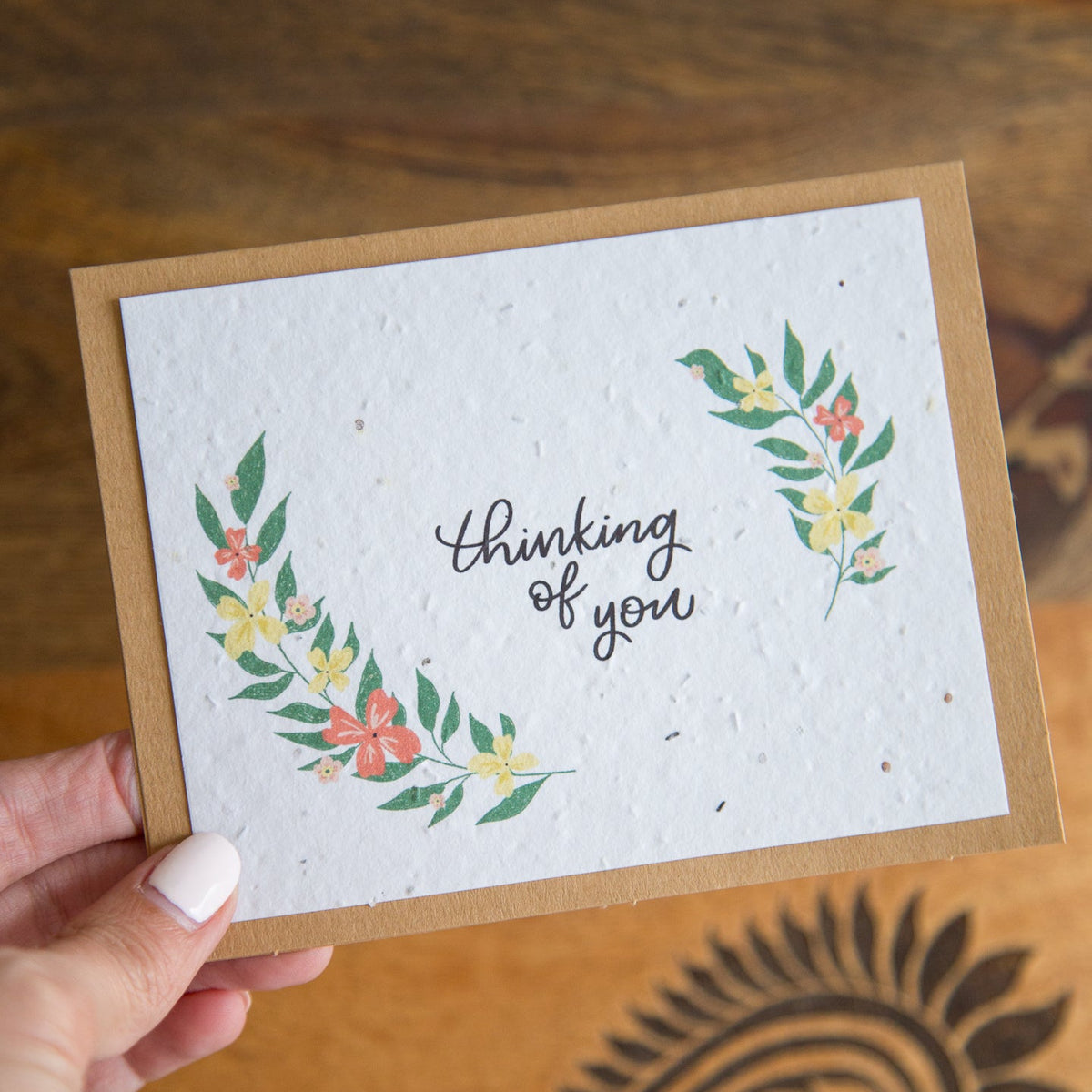 A handmade greeting card with a kraft paper border and a white textured center featuring the phrase 'thinking of you' in black script, decorated with green leafy wreaths and small pink and yellow flowers.