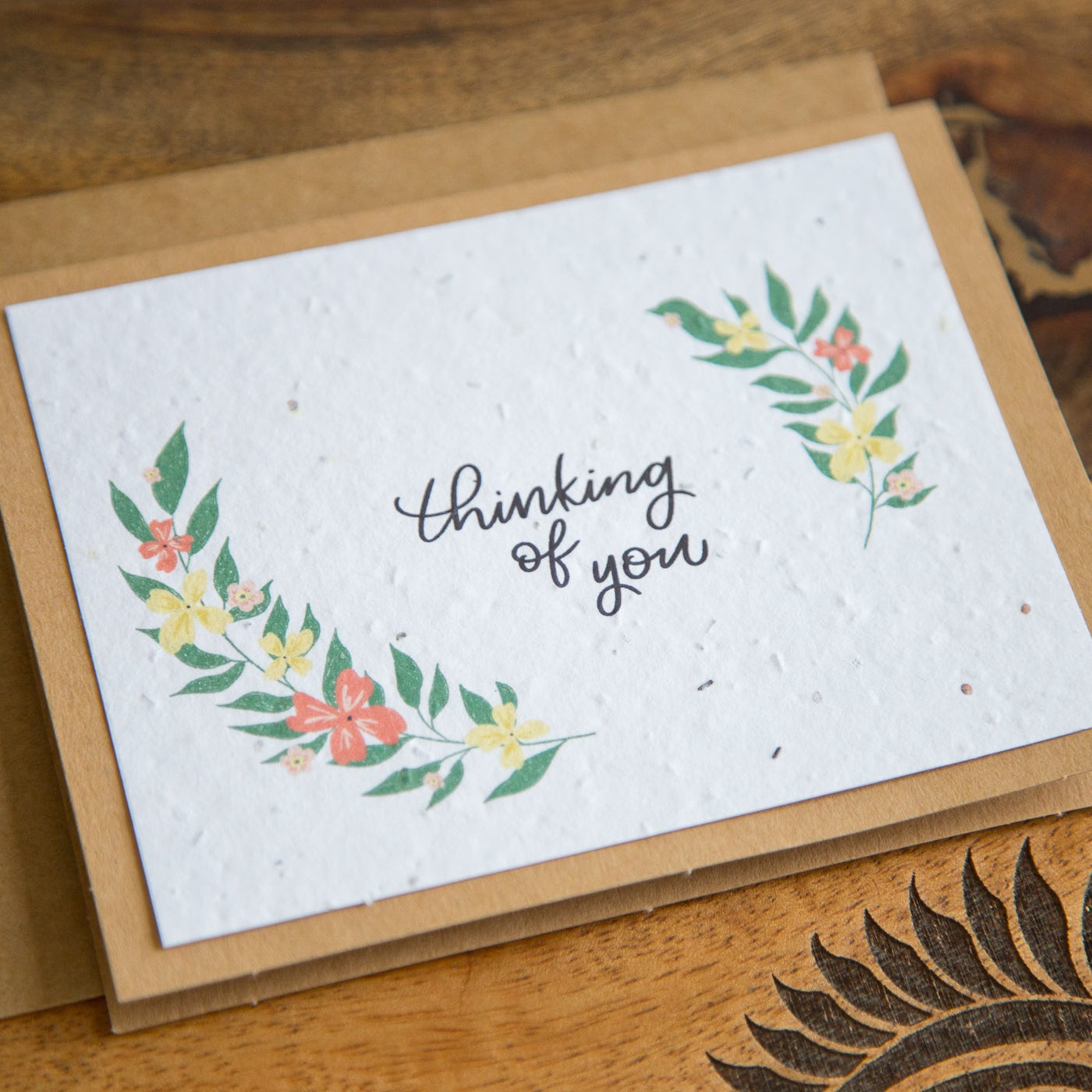 A square white textured greeting card with the phrase 'thinking of you' in black script, decorated with green leaves and small red and yellow flowers, resting on a brown kraft envelope over a wooden surface.