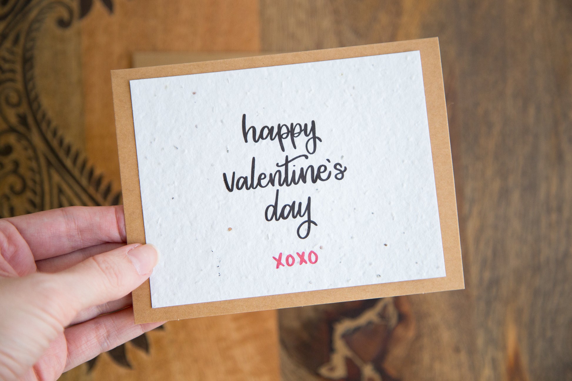 A hand holding a plantable greeting card with the words "happy Valentine's Day xoxo" printed in calligraphy in black on white paper