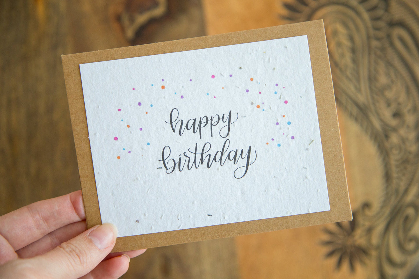 A hand holding an A2 sized greeting card printed on plantable seed paper with the words "happy birthday" and coloured confetti