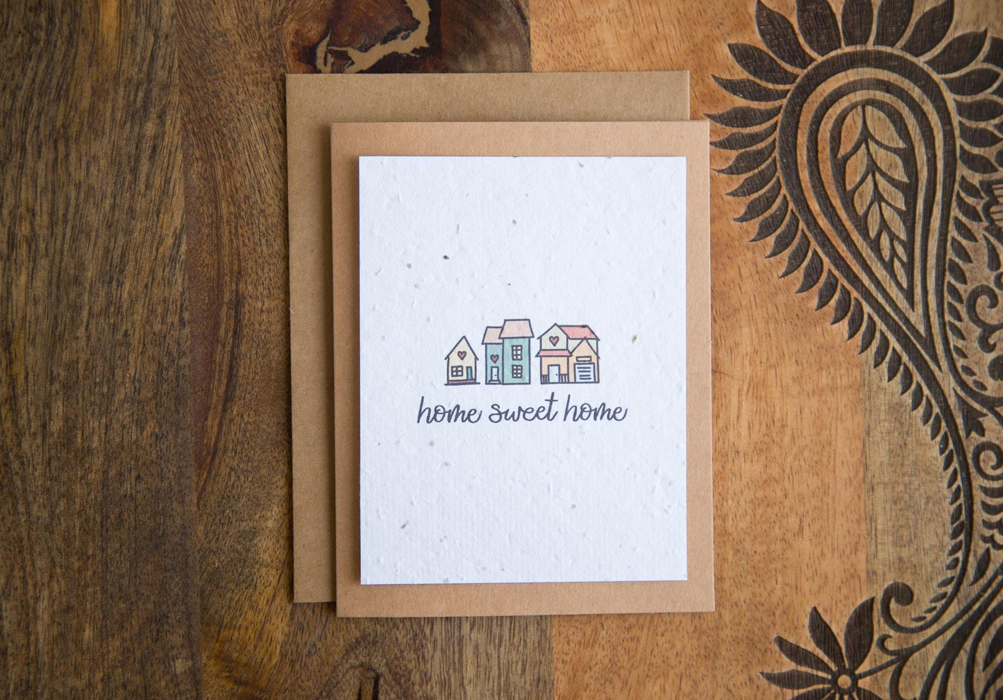 A2 greeting card on a table with the words "home sweet home" and an illustration of 3 small houses printed on white seed paper 