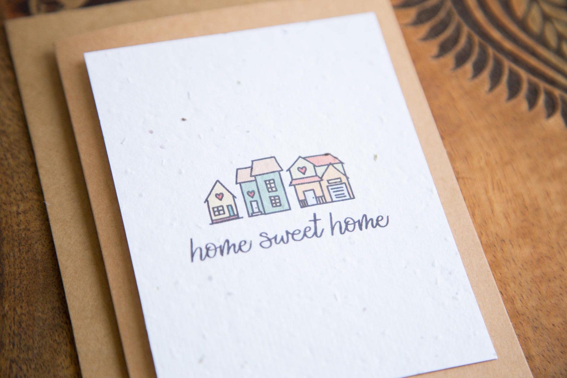 A2 greeting card on a table with the words "home sweet home" and an illustration of 3 small houses printed on white seed paper 