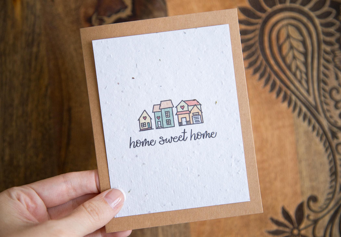 A hand holding an A2 greeting card with the words "home sweet home" and an illustration of 3 small houses printed on white seed paper 