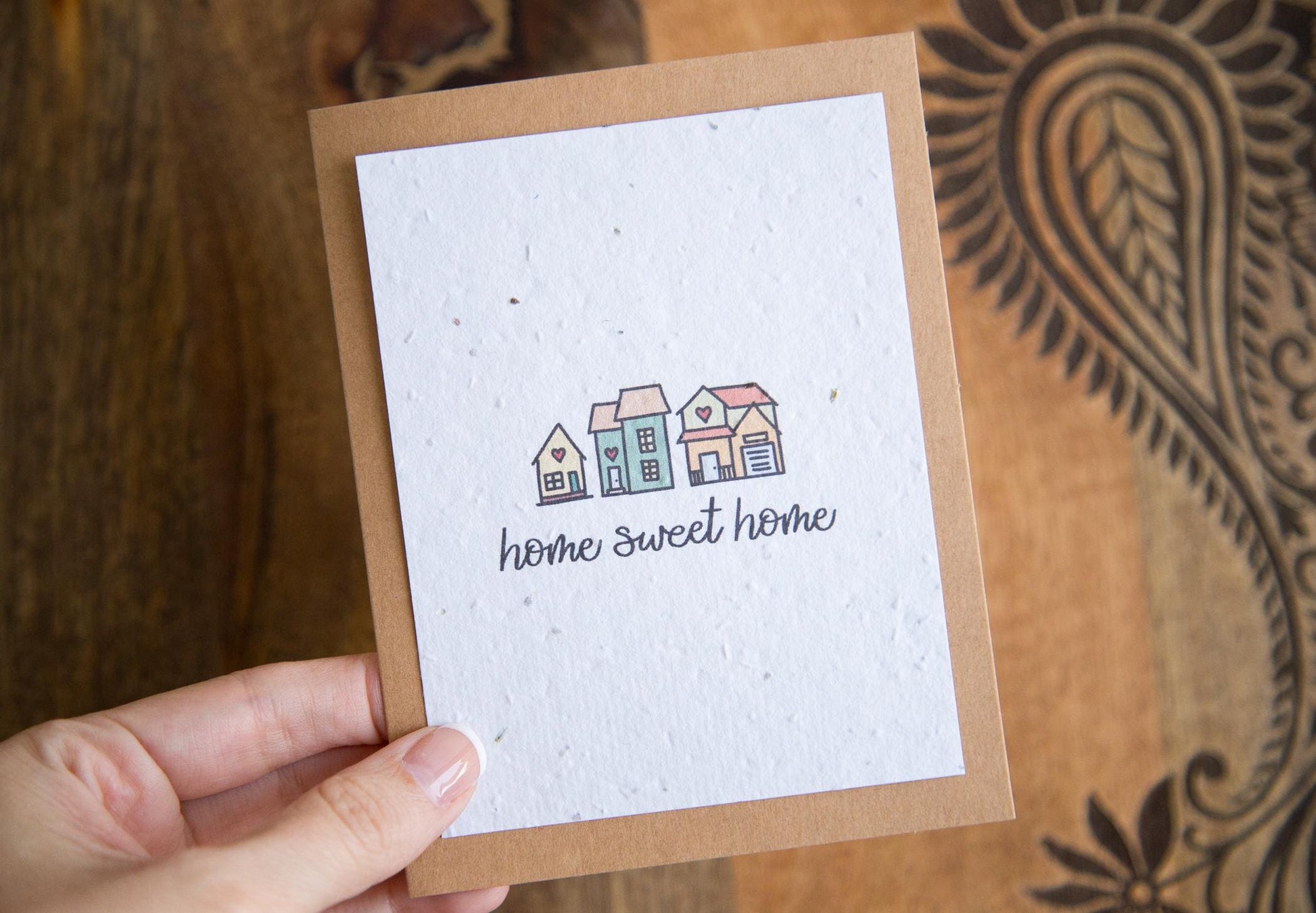 A hand holding an A2 greeting card with the words "home sweet home" and an illustration of 3 small houses printed on white seed paper 