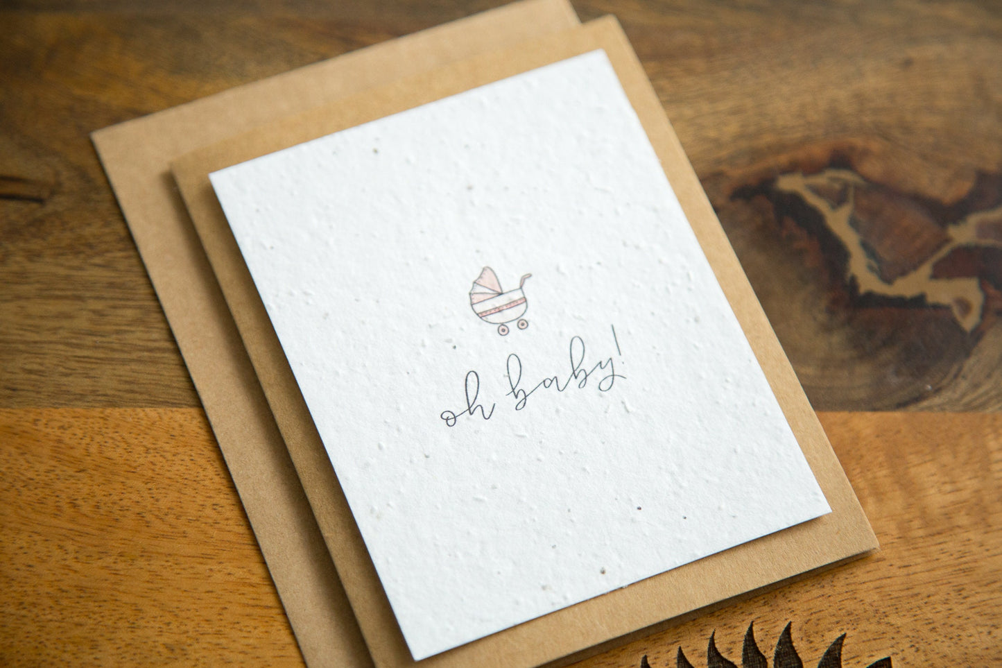A2 greeting card on a table with the word "oh baby!" and an illustration of a baby carriage in pink printed on white seed paper 