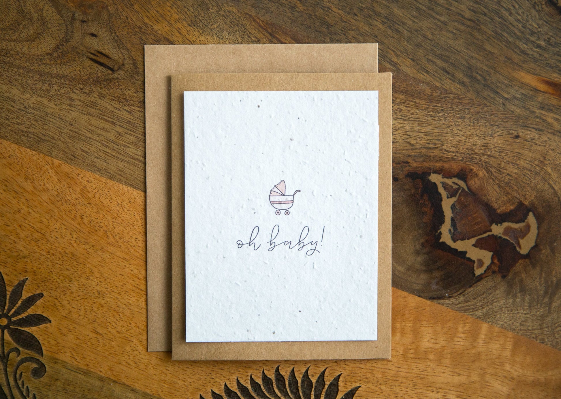 A2 greeting card on a table with the word "oh baby!" and an illustration of a baby carriage in pink printed on white seed paper 