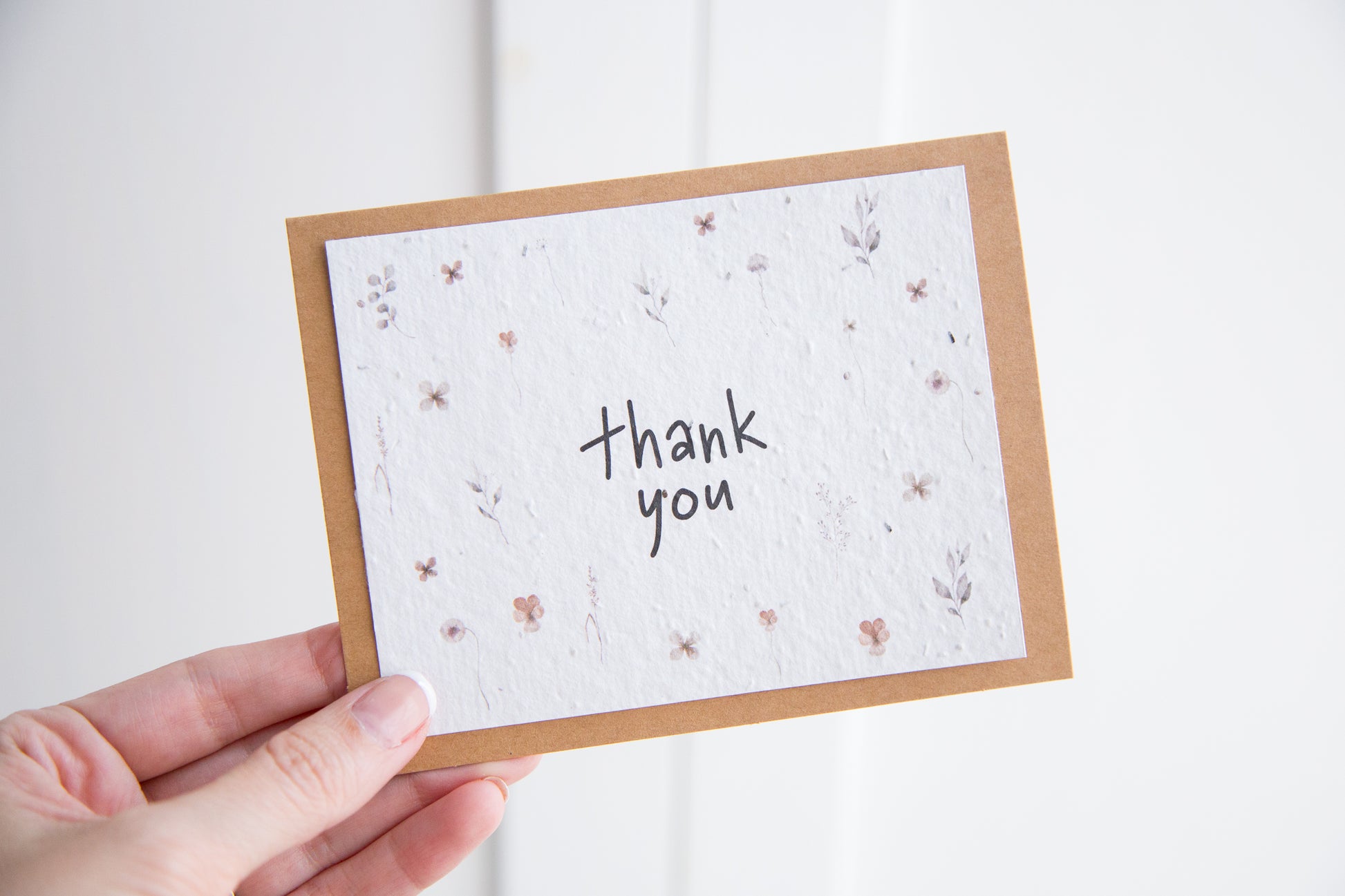 A hand holding an A2 greeting card with the words "thank you" surrounded by watercolour florals printed on white seed paper 
