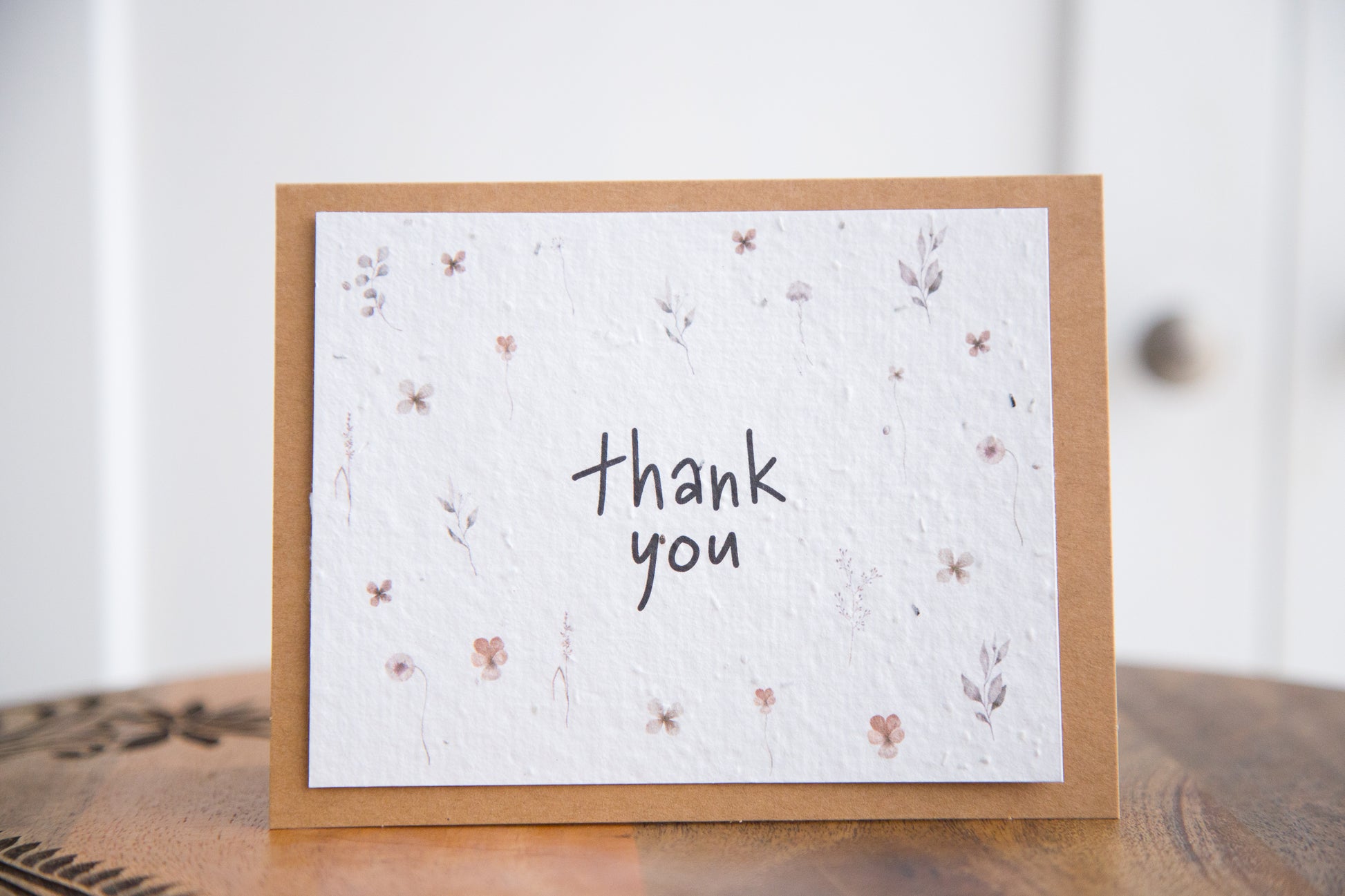 An A2 greeting card on a wooden table with the words "thank you" surrounded by watercolour florals printed on white seed paper 